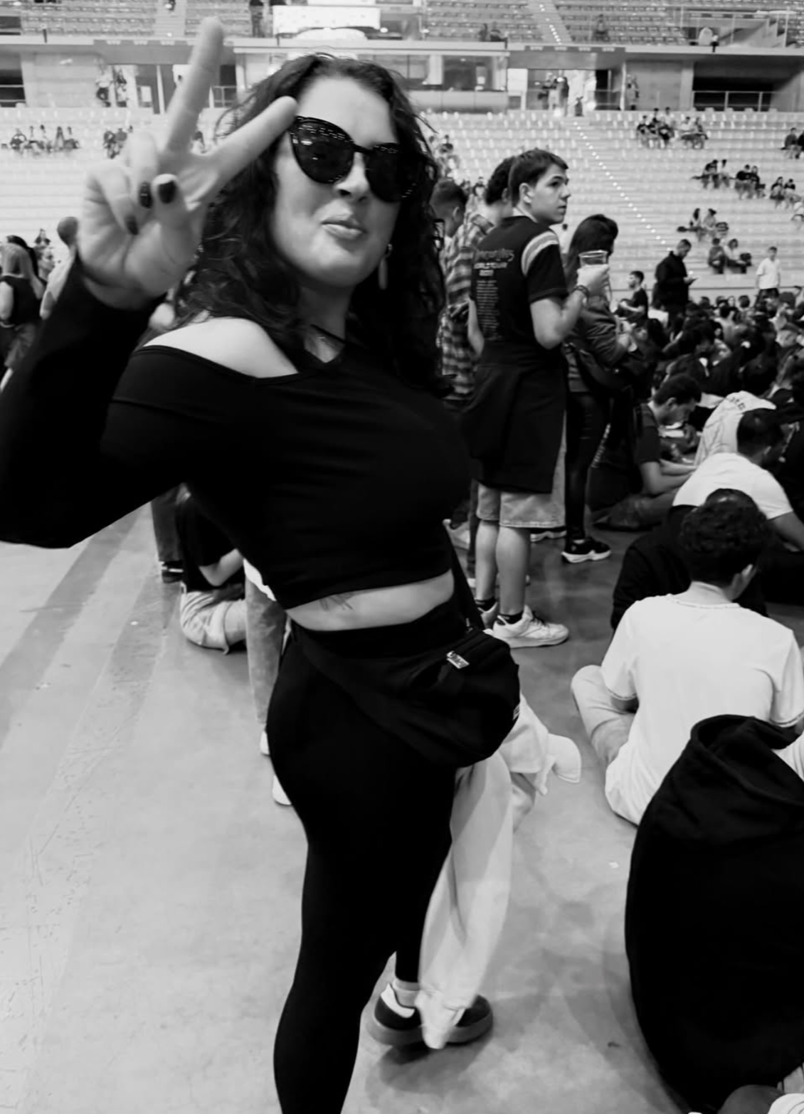 A woman with curly hair, sunglasses, and a black off-shoulder top making a peace sign at a crowd event in an indoor stadium.