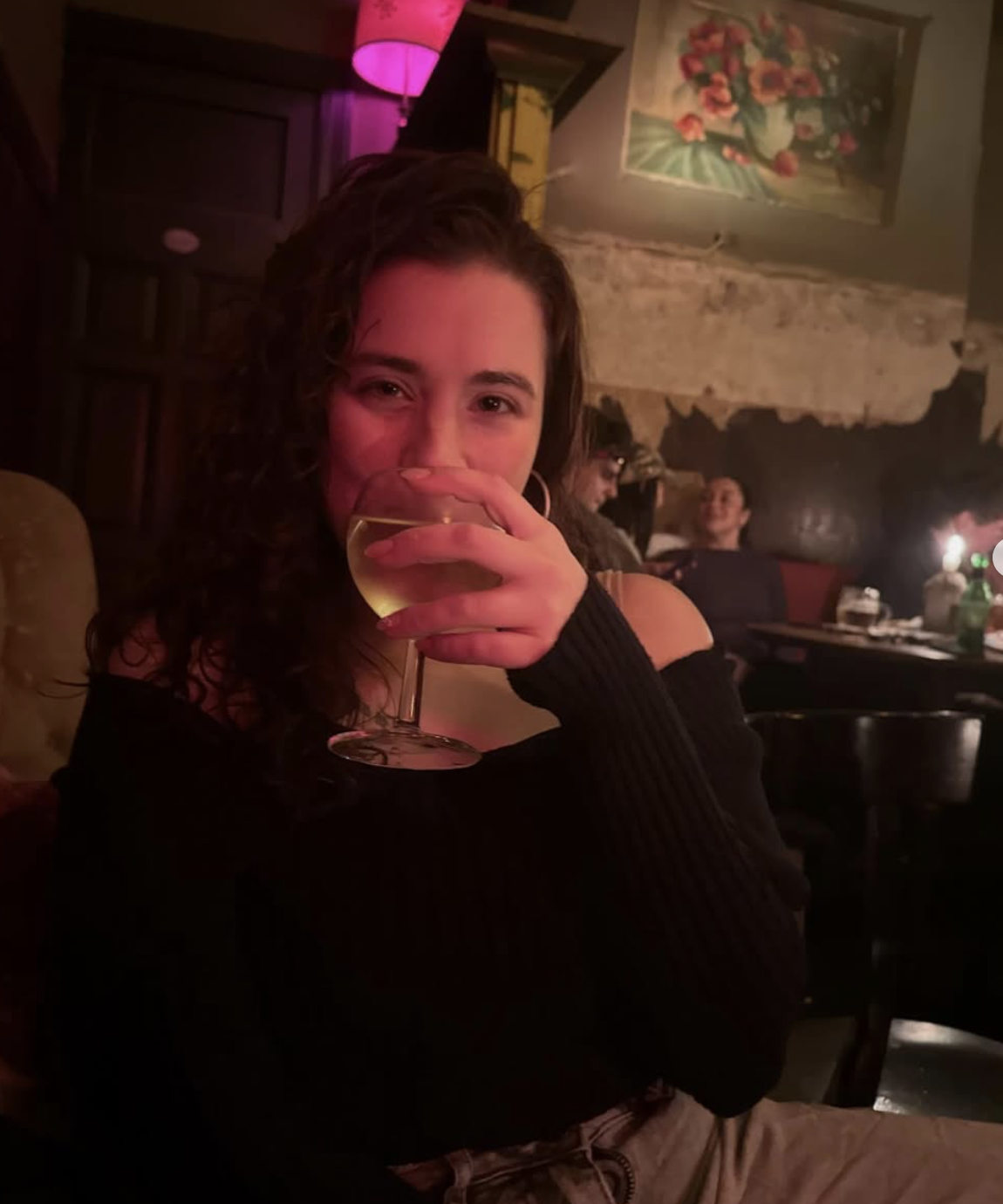 A young woman with curly hair in a black off-the-shoulder top sitting in a dimly lit restaurant or bar, smiling and holding a glass of white wine in front of her face. The background shows a stone wall, paintings, and other patrons at tables.