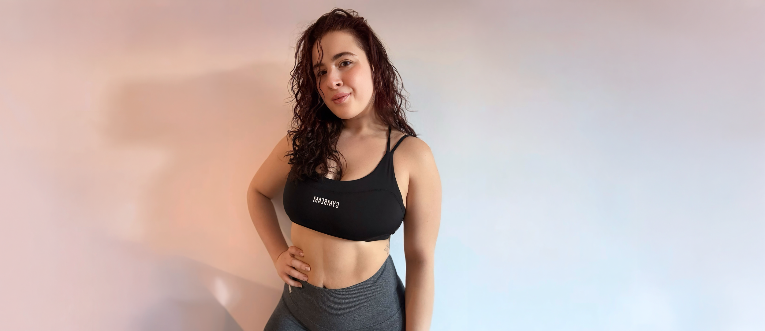 Young woman with curly brown hair posing in athletic wear against a plain wall, wearing a black sports bra with the word 'GYMBEAM' on it and gray leggings.