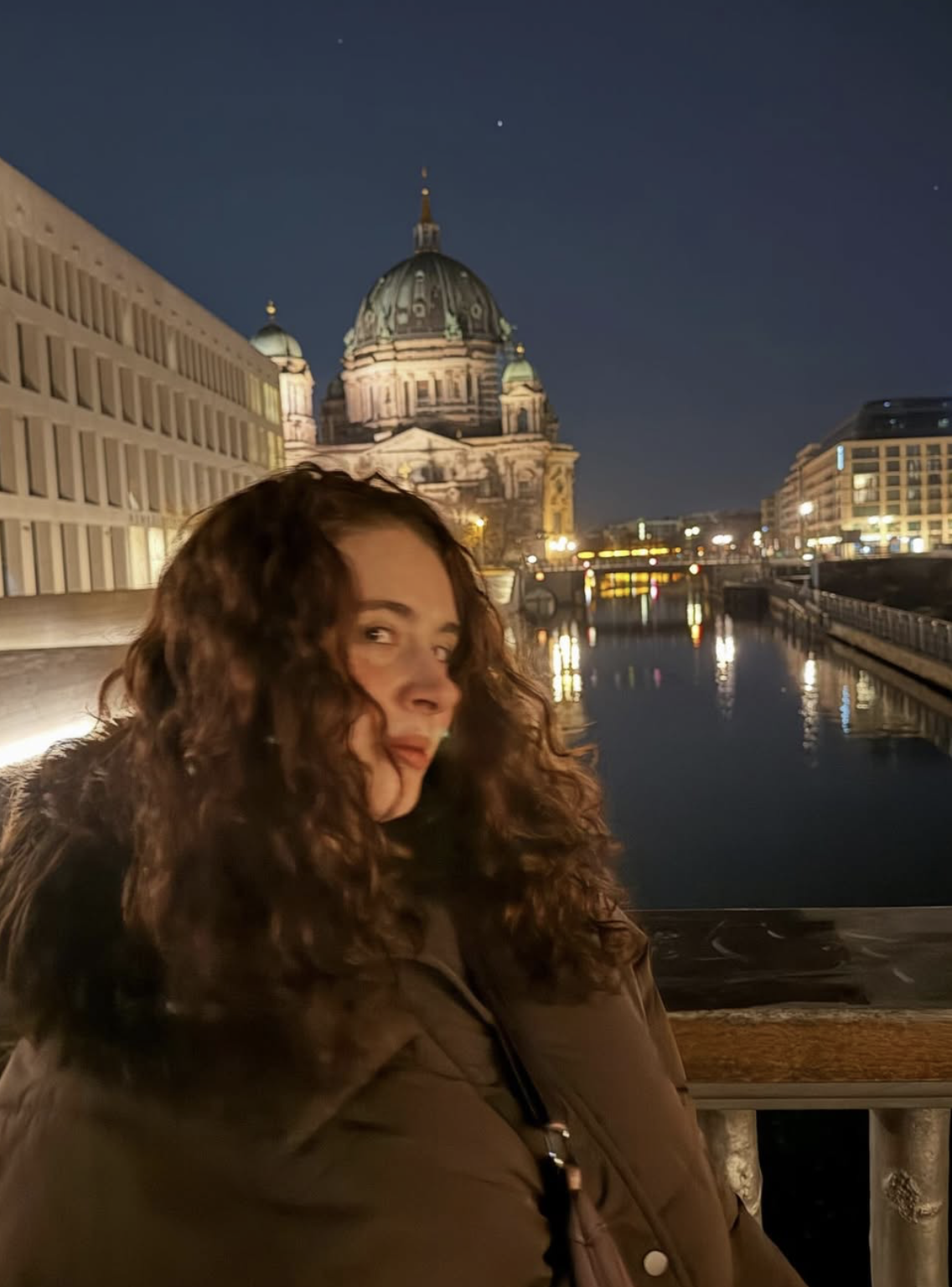 A woman with curly hair in a brown jacket standing by a canal at night, with a domed historic building and city lights reflected in the water behind her.