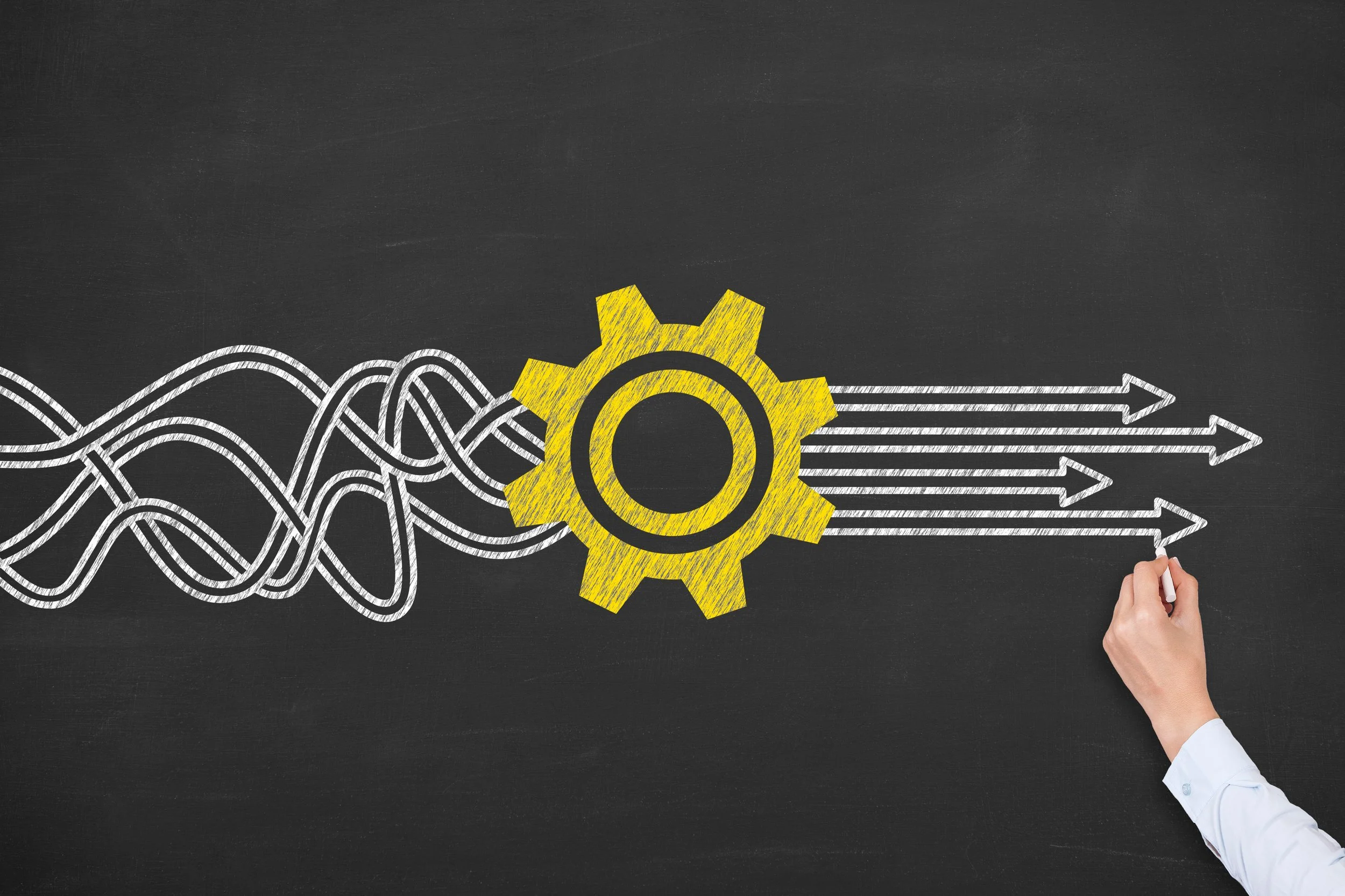 Drawing of a yellow gear with white lines and arrows representing interconnected processes or systems, on a blackboard background, with a hand holding chalk in the bottom right corner.