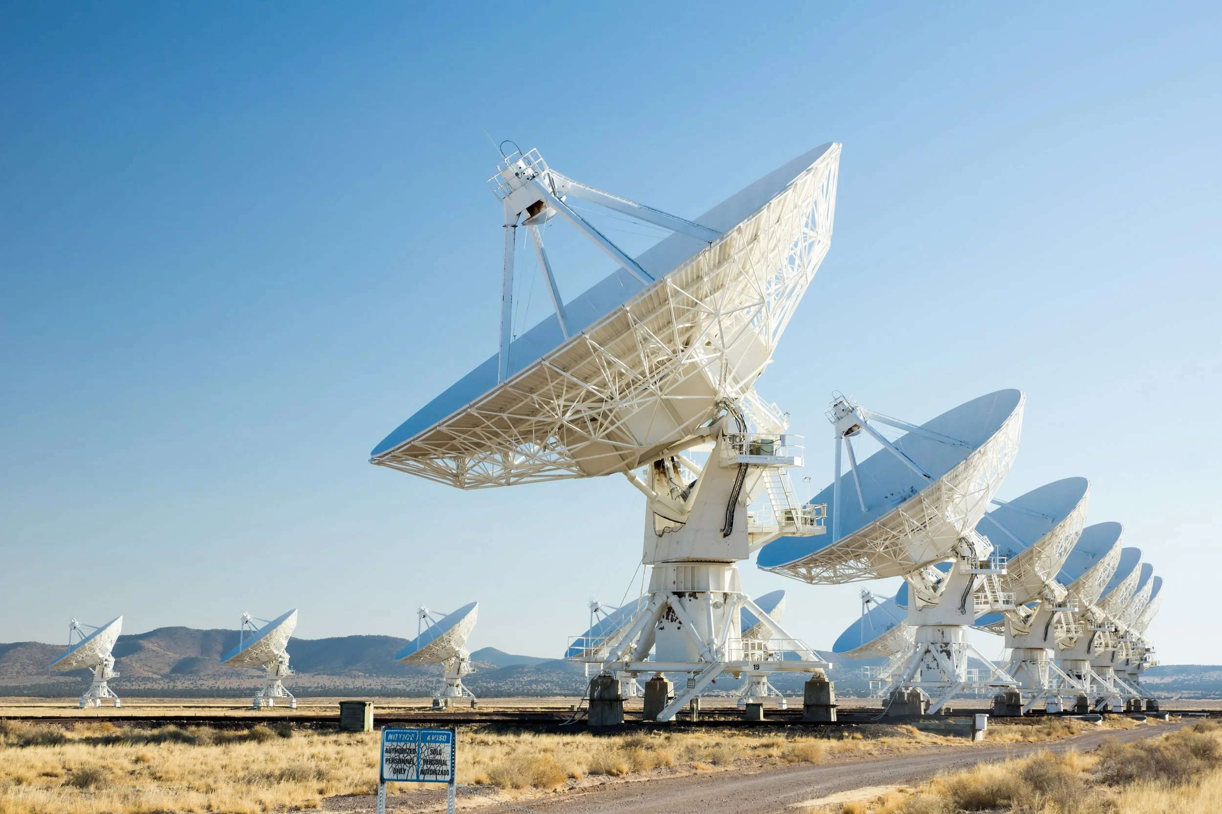 Multiple radar dish antennas lined up outdoors in a desert landscape under a clear blue sky.