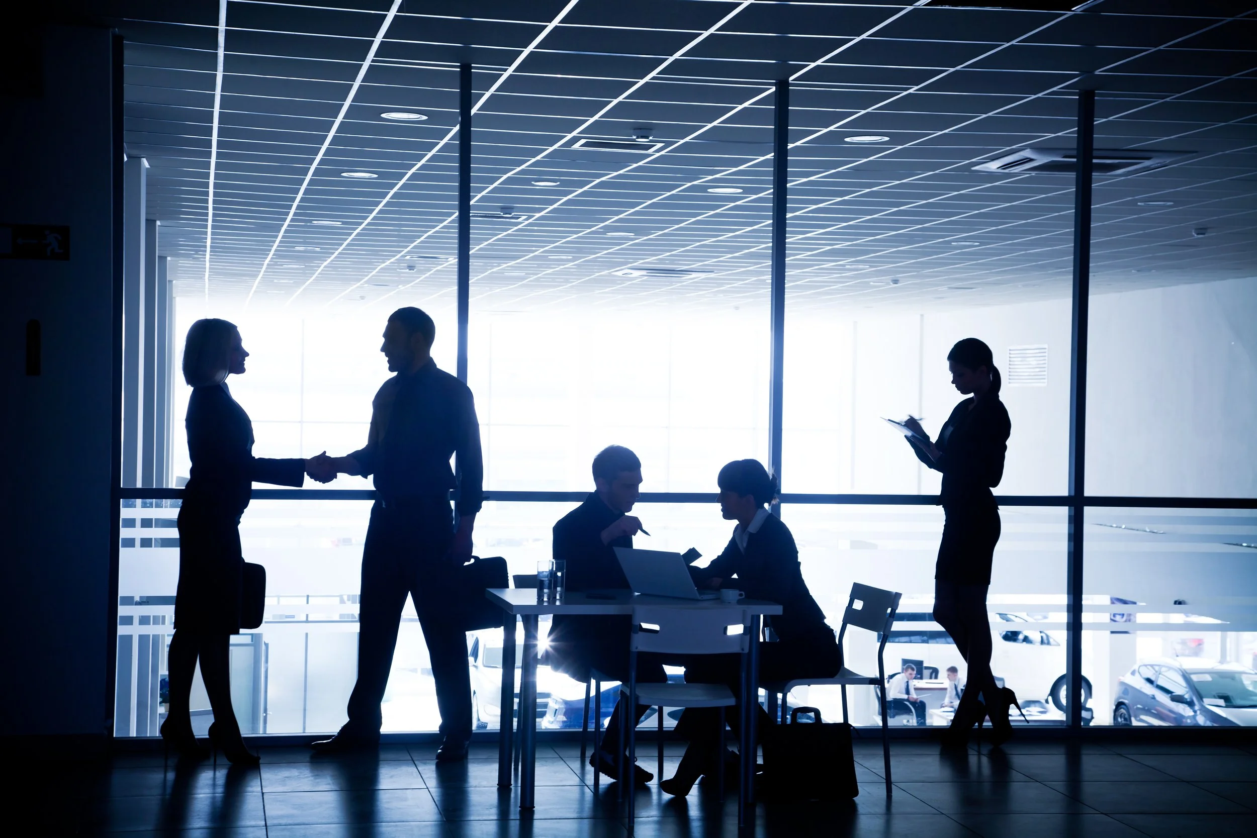 Silhouettes of businesspeople engaging in conversations and working on tablets in a modern, glass-walled office with bright natural lighting.