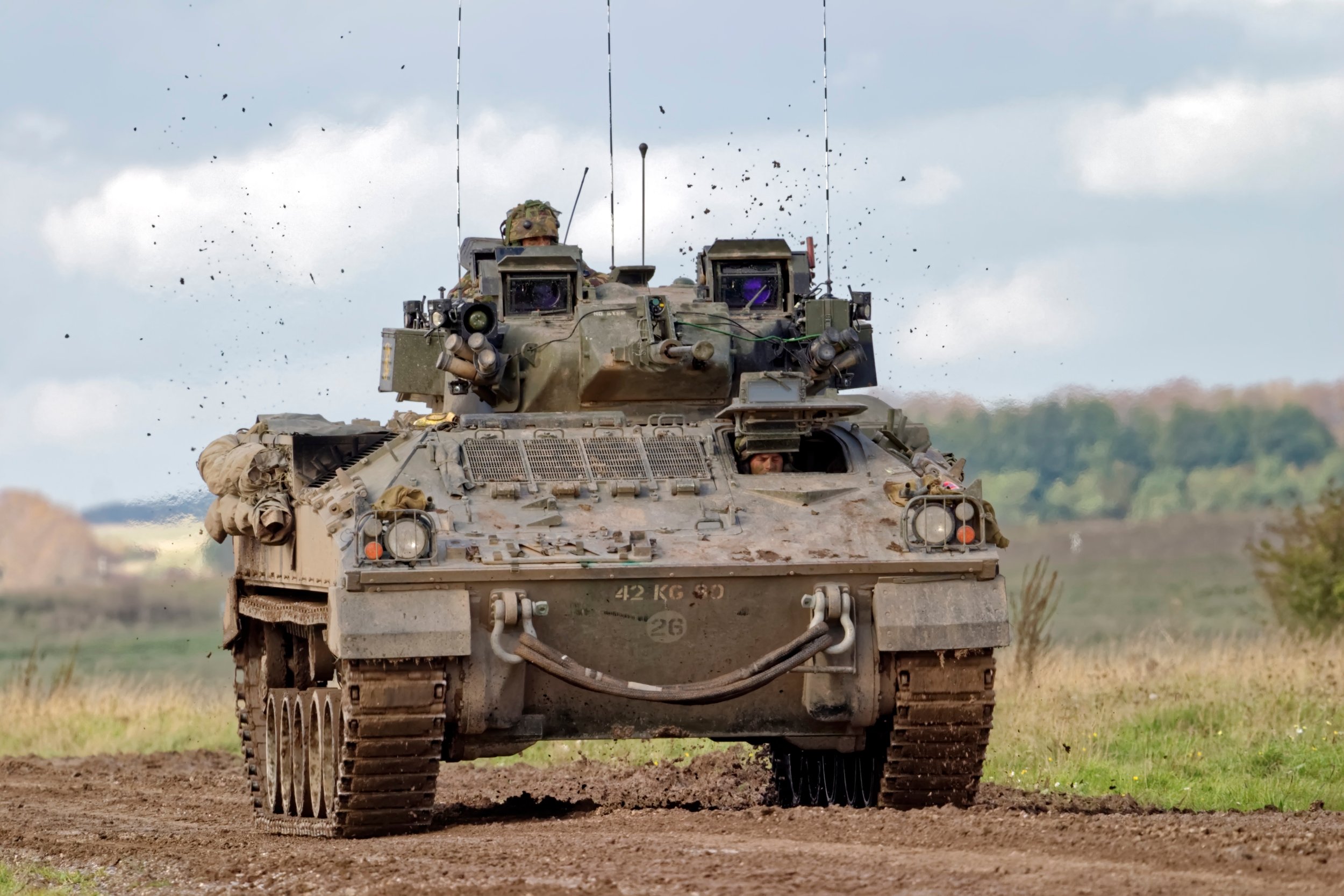 A military tank moving on a dirt road with a soldier visible through the turret window. The tank is kicking up dirt and debris as it travels, under a cloudy sky.