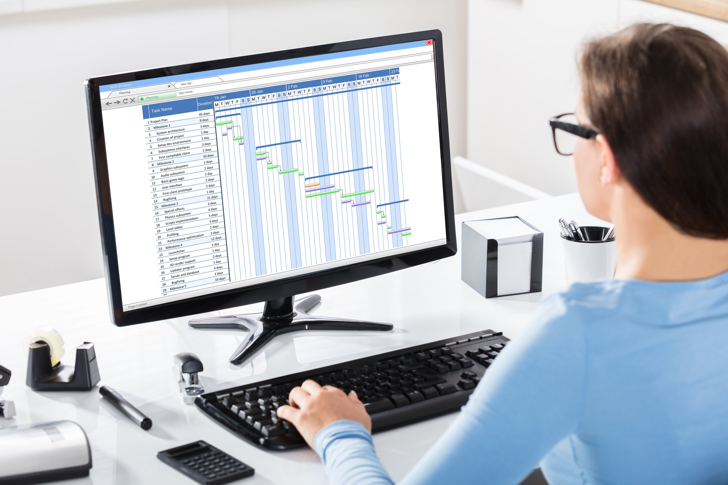 A person working at a computer resembling a project management Gantt chart with tasks, durations, and timelines displayed on the screen.