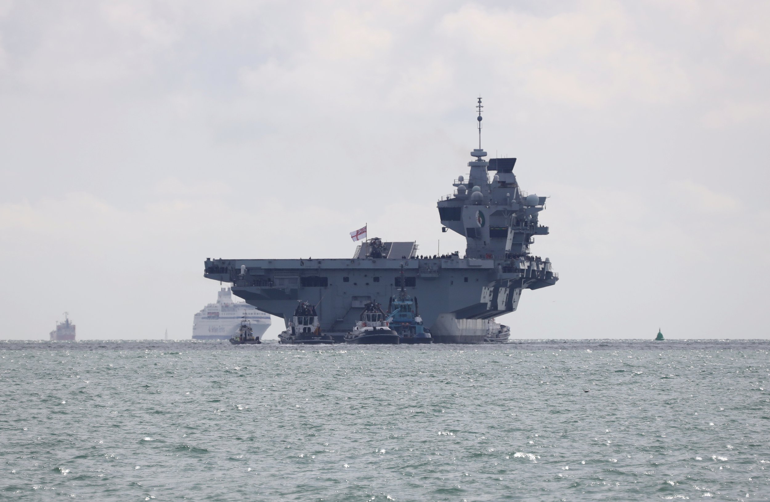 A large aircraft carrier submarine being towed by several tugboats on the water with ships visible in the background.