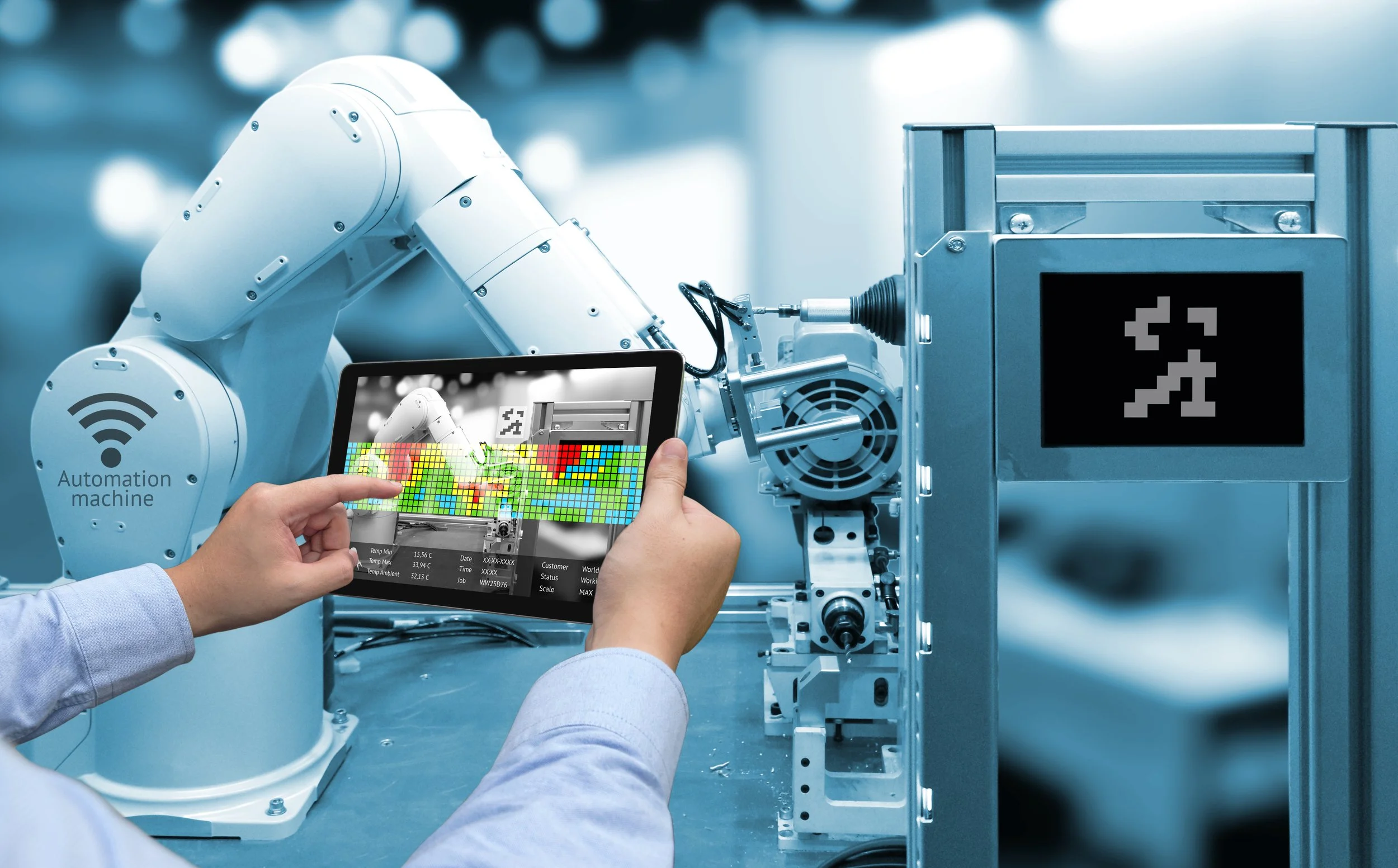 A person in technical clothing operating a robot arm in an industrial setting. The person holds a tablet displaying a thermal or heat map overlay of the robot, with data and metrics.
