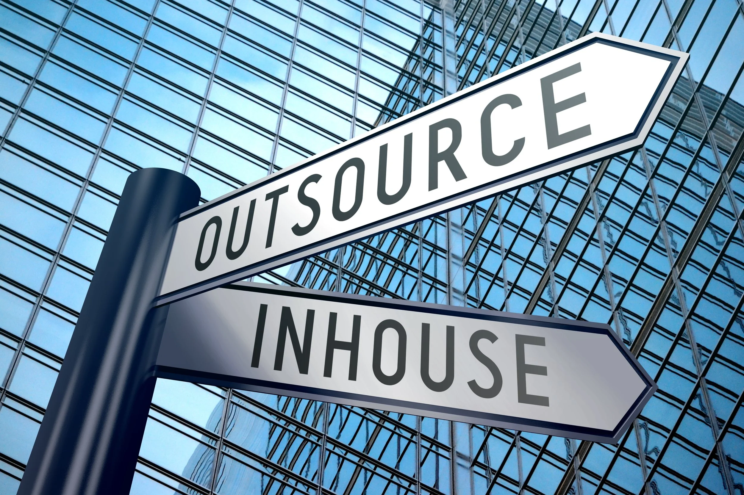 Street signs with directions, including 'OUTSOURCE' and 'INHOUSE', mounted on a pole in front of a modern glass building.