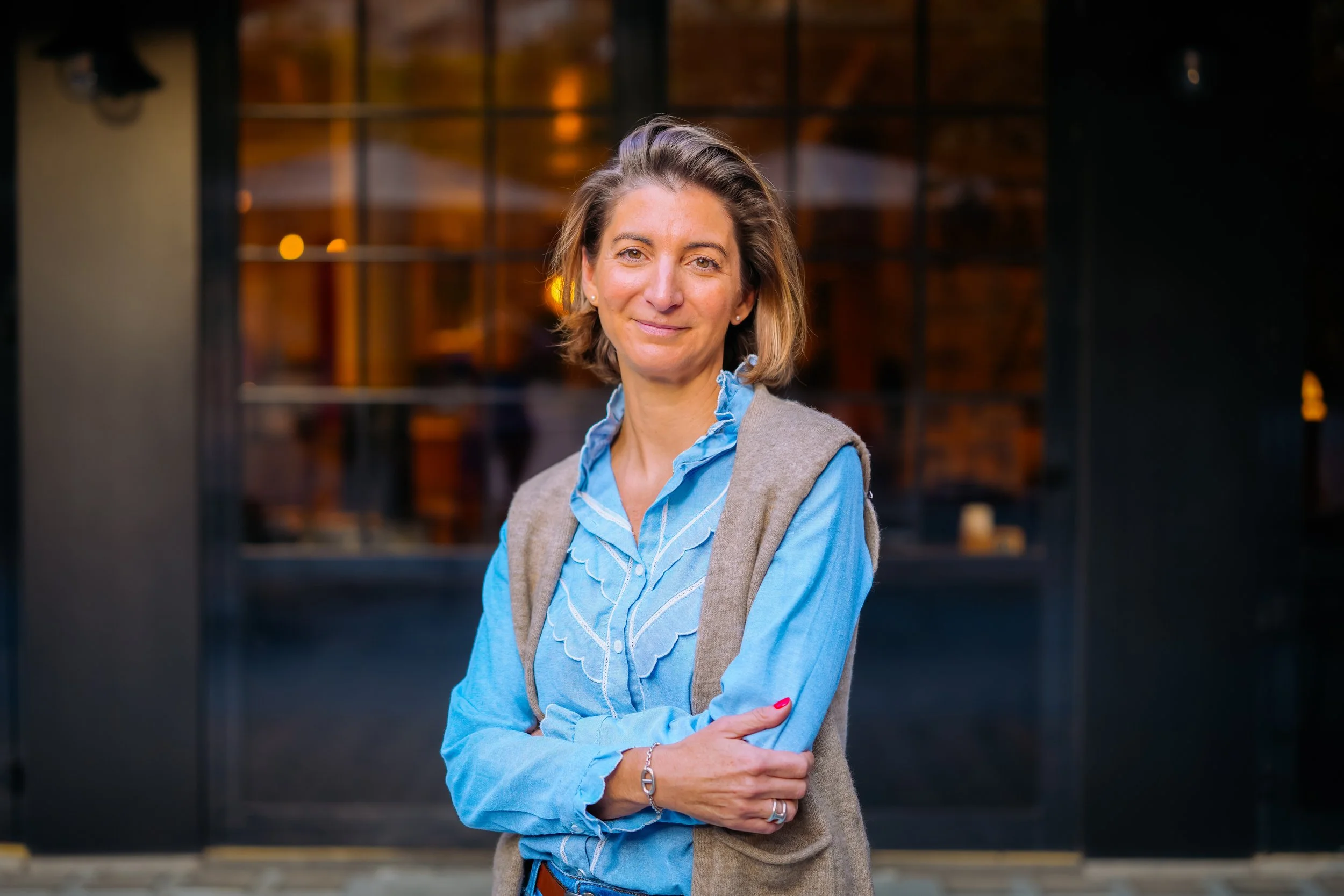 Femme souriante portant un chemisier bleu avec une veste beige, debout devant une porte en verre dans un cadre urbain.