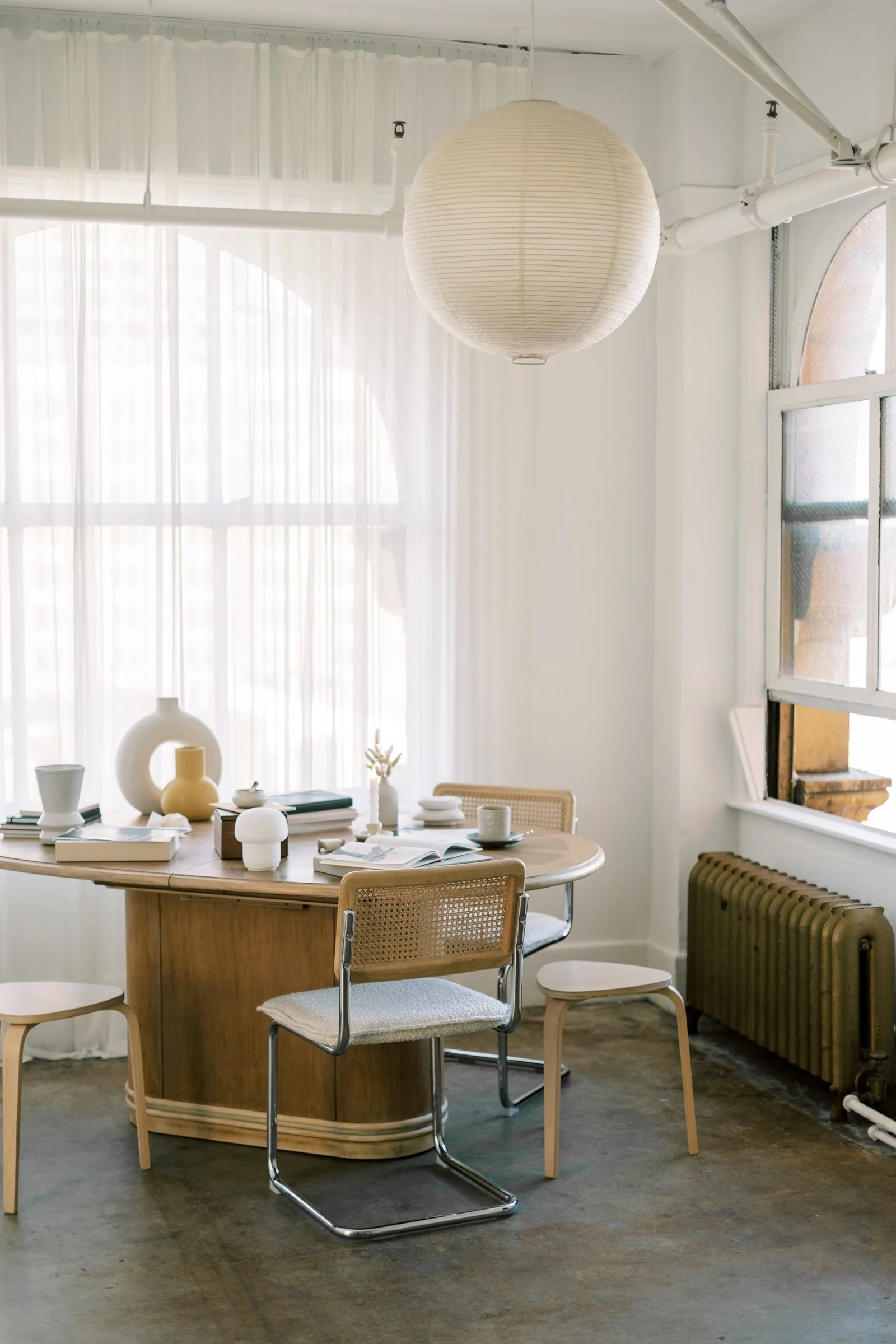 Une salle lumineuse avec une table en bois, plusieurs chaises, vases décoratifs, livres, une tasse et un journal. Les murs sont blancs, avec de grandes fenêtres laissant passer la lumière.
