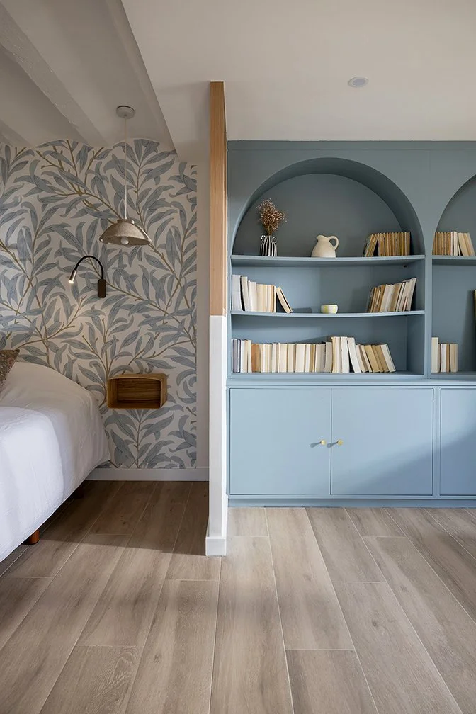 Une chambre avec un lit blanc et un mur décoratif avec un motif de feuillage, séparée d'une bibliothèque bleue contenant des livres et décorations, sur un sol en bois clair.