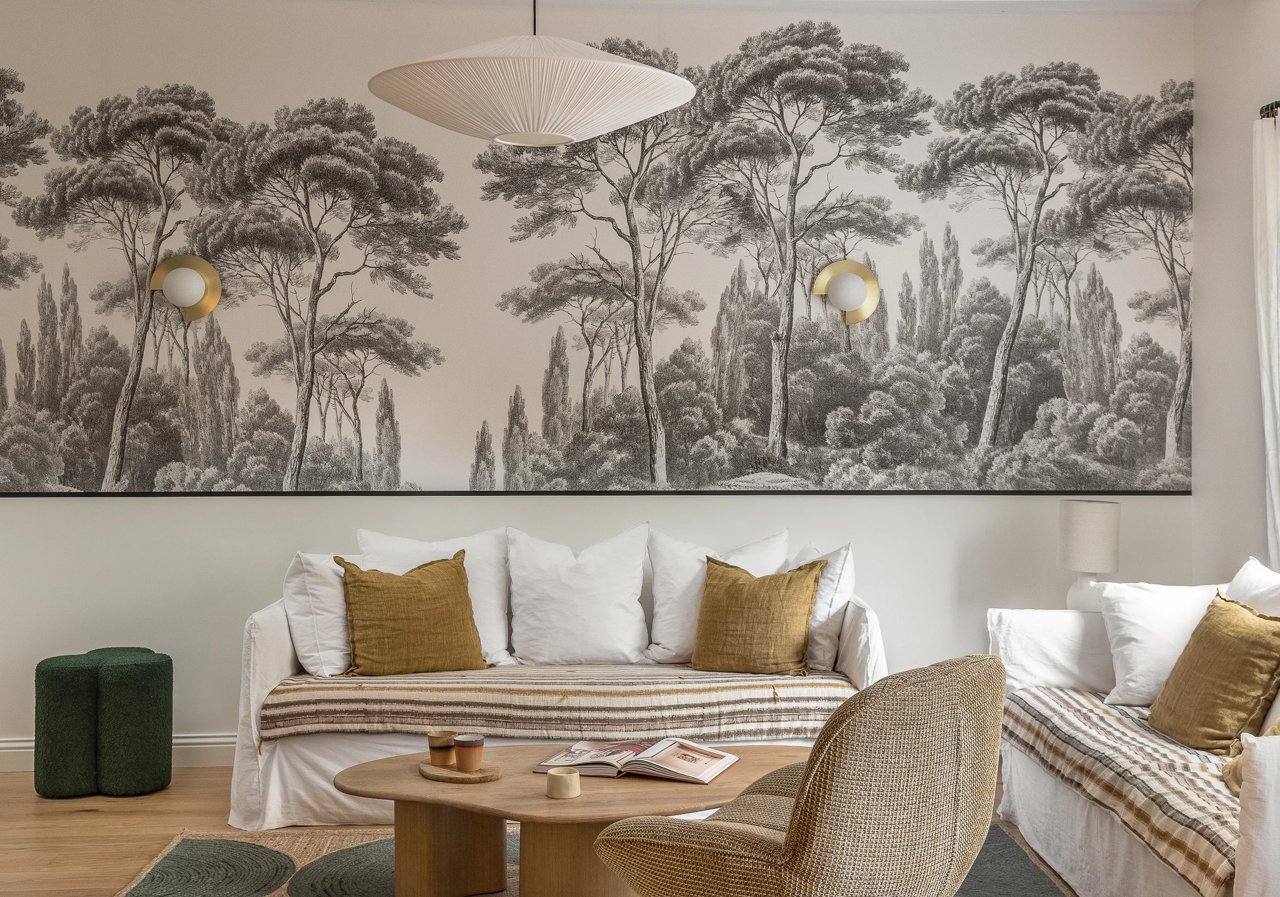 Un salon avec un grand mural en noir et blanc représentant des arbres, deux canapés blancs avec des coussins marron, une table basse en bois avec des livres et des tasses, un fauteuil beige, une lampe de table blanche, et un plafonnier blanc