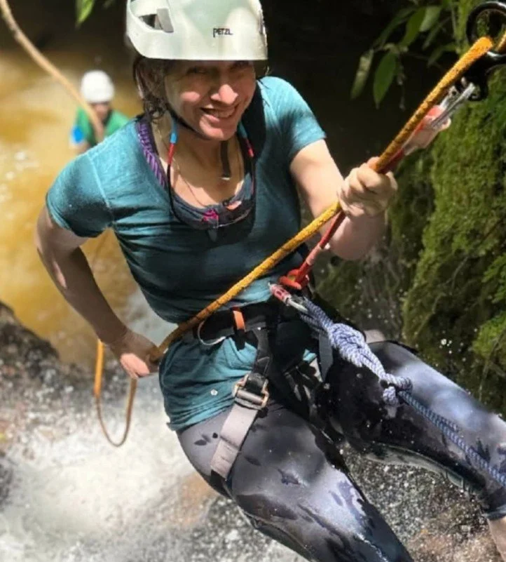 Person wearing a helmet and harness, climbing or rappelling beside a moss-covered rock face, with water splashing around.