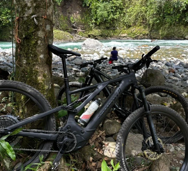 Two black electric mountain bikes leaning against a tree by a rocky riverbank. A person is sitting on the rocks near the river, surrounded by dense green forest.