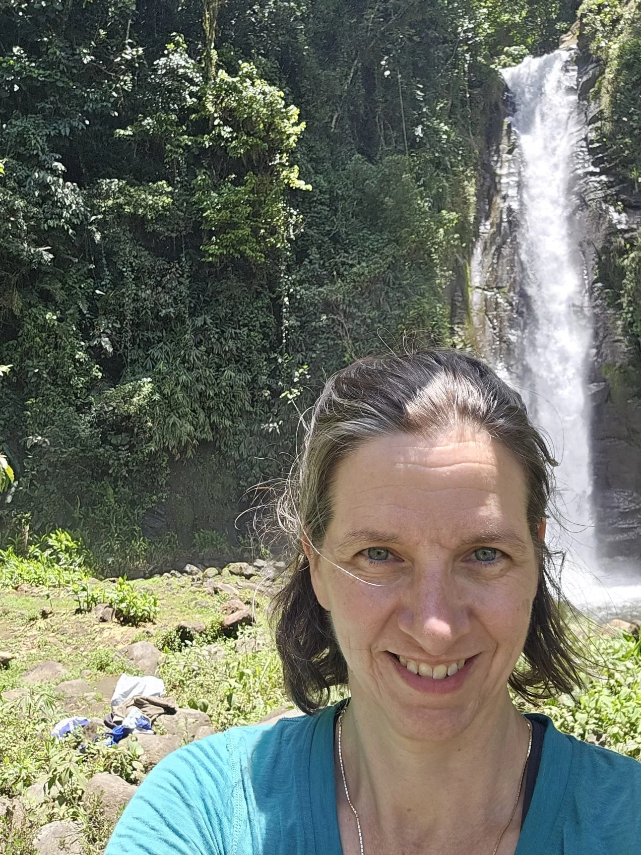 A woman smiling in front of a waterfall surrounded by lush greenery.