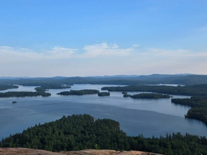 Aerial view of a large body of water with numerous small islands and surrounding forested land under a partly cloudy sky.