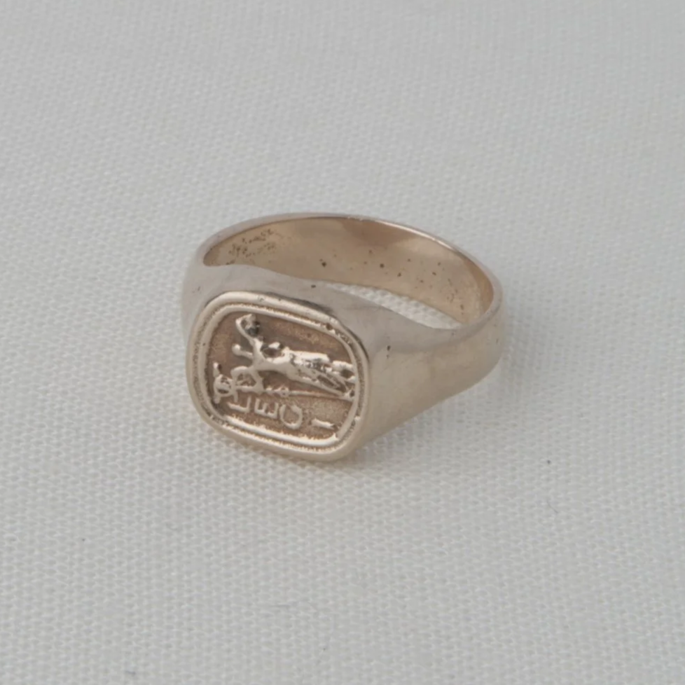 A silver ring with an engraved crest or emblem on top, placed on a white textured background.