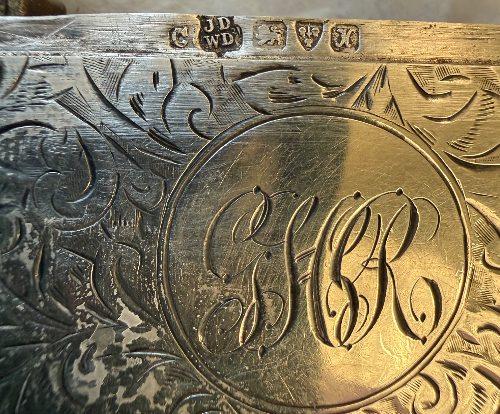 Close-up of engraved silver Vesta case with decorative patterns and initials 'G' and 'L' inside a circle.