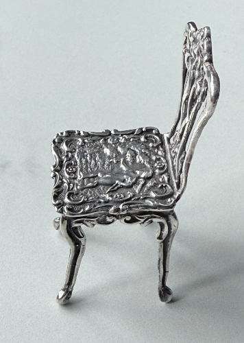 Small, ornate silver chair with intricate carvings and classical figures on the seatback.