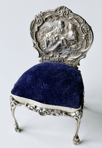 An ornate silver miniature chair with a decorative backrest depicting a classical scene, covered with a dark blue velvet cushion.