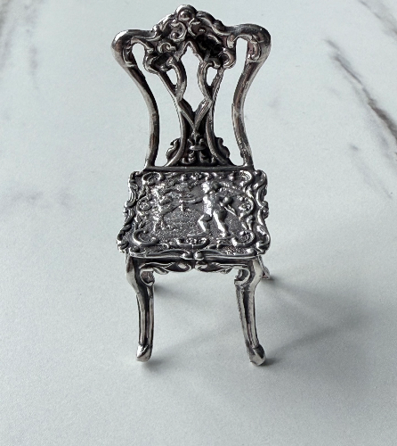 Small ornate silver chair figurine with detailed carvings, placed on a white surface.