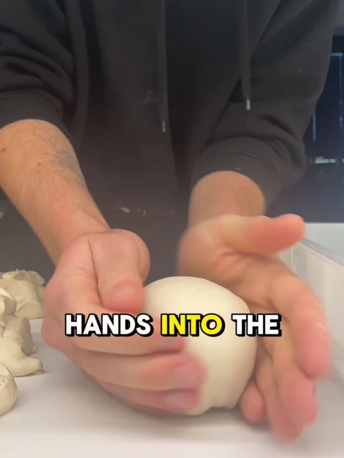 A person kneading dough with their hands, with the text 'HANDS INTO THE' overlaid. The person is wearing a black hoodie, and there are small pieces of dough on the white surface.