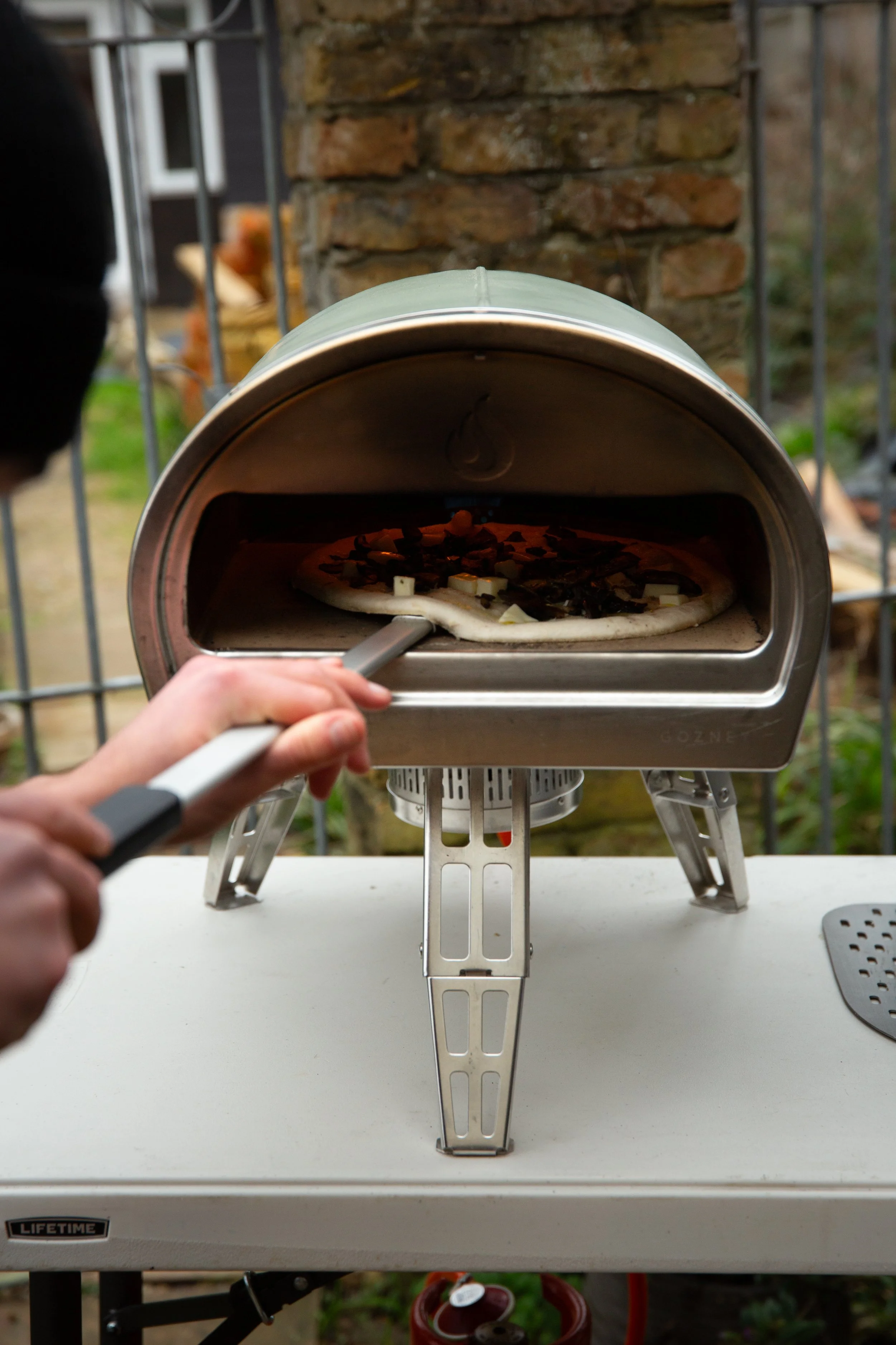 A person is placing a pizza into a small, stainless steel outdoor pizza oven, with a brick wall and a garden in the background.