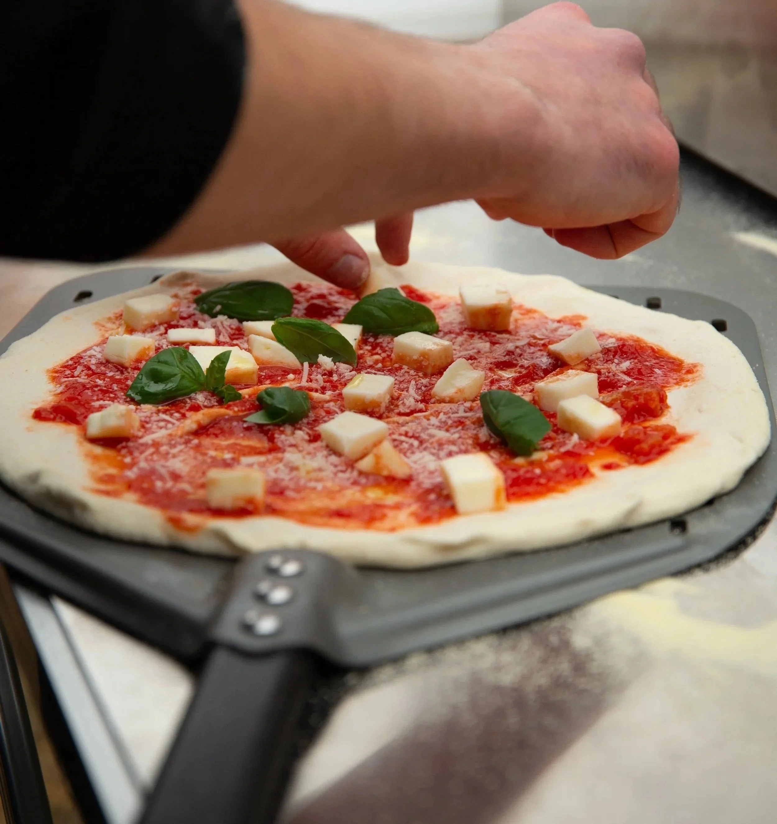 Person placing fresh basil leaves and cheese cubes on a pizza with tomato sauce and an unbaked crust.