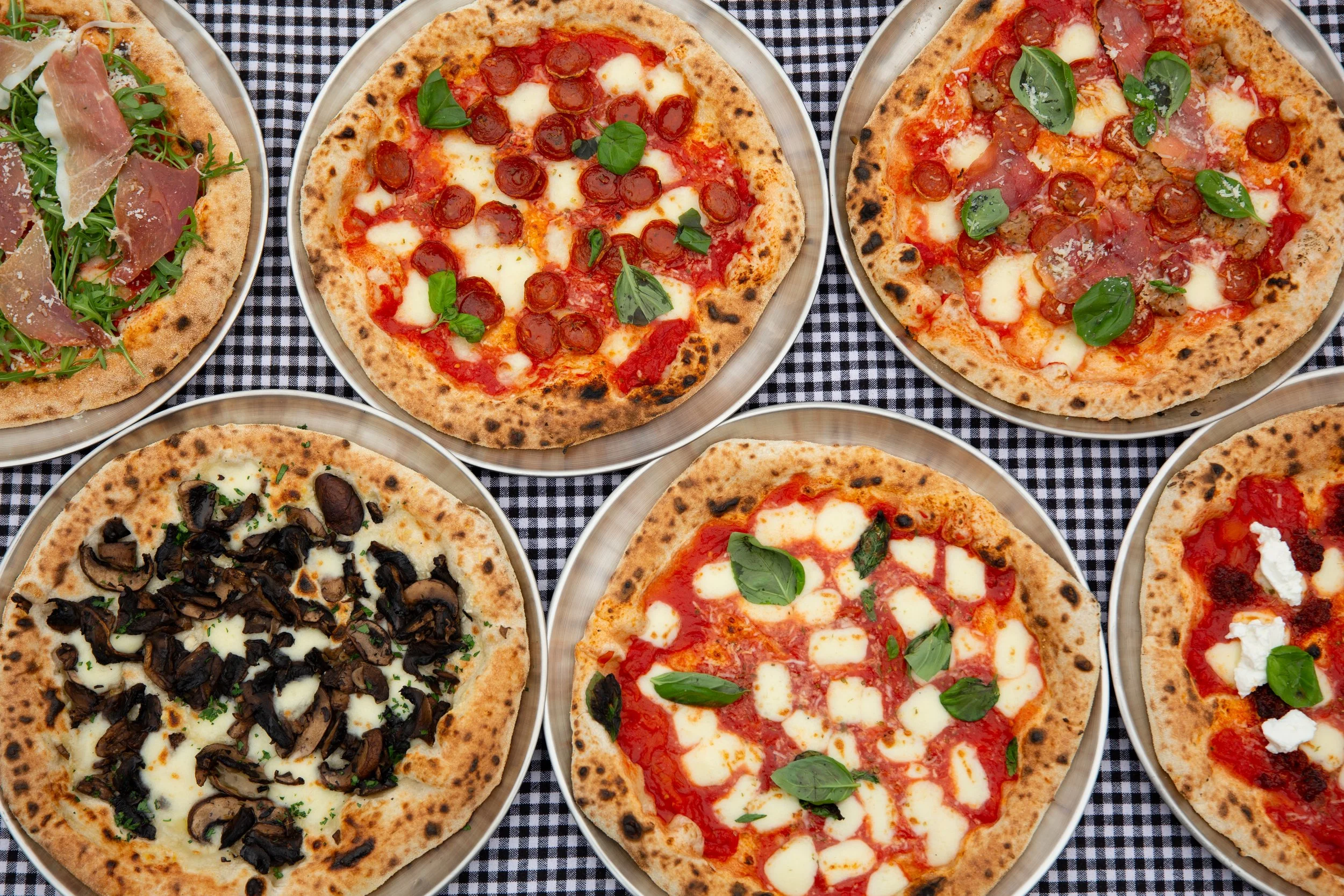 Six pizzas with various toppings on metal trays placed on a black and white checkered tablecloth.