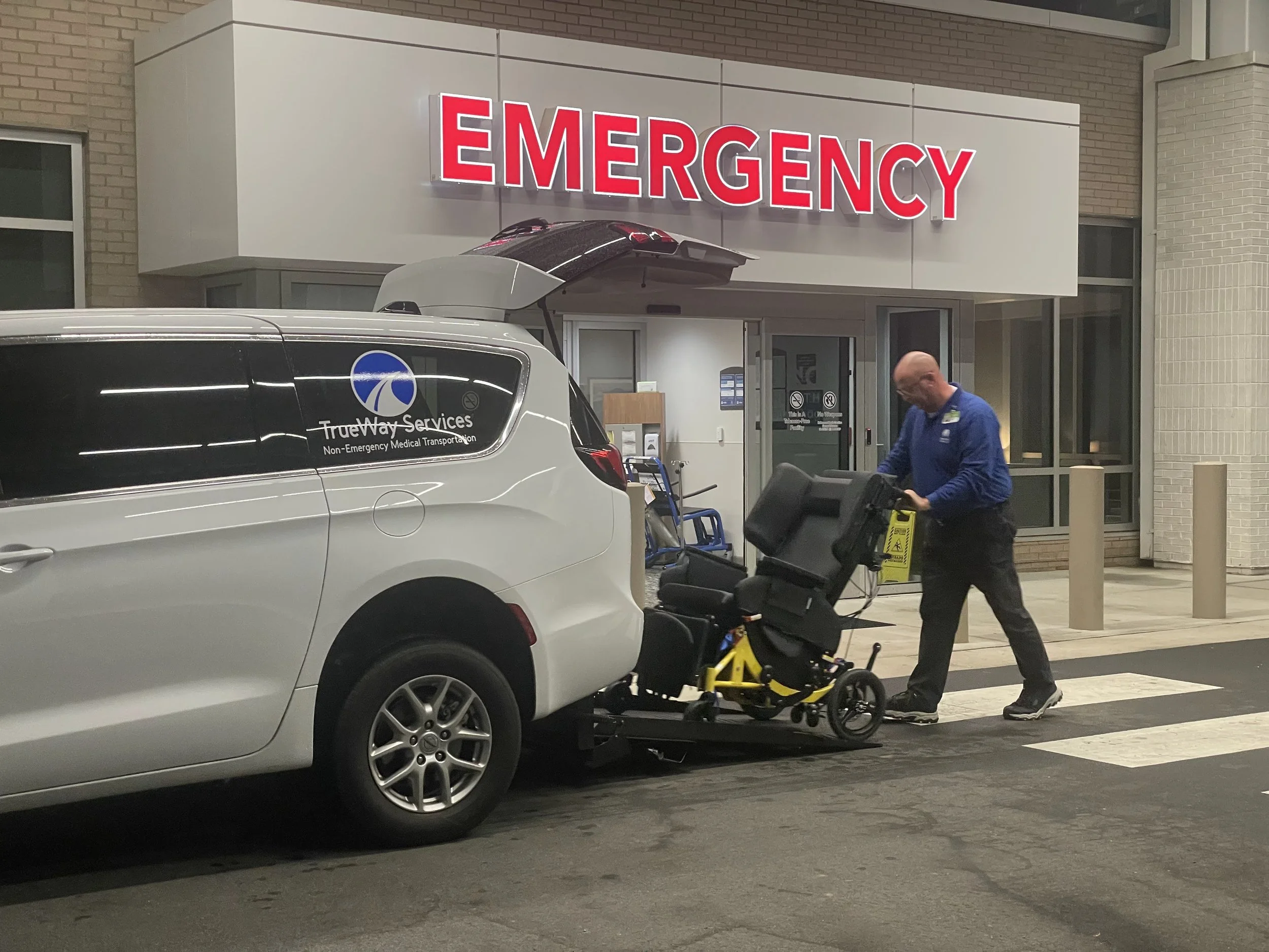 A person in a blue jacket is pushing a wheelchair into the back of an ambulance at a hospital emergency entrance. The entrance has a large red illuminated sign that reads 'EMERGENCY', and there is a white medical service vehicle labeled 'TrueWay Services Non-Emergency Medical Transportation' parked nearby. The scene is taken inside a well-lit urban hospital area with brick walls and glass doors.