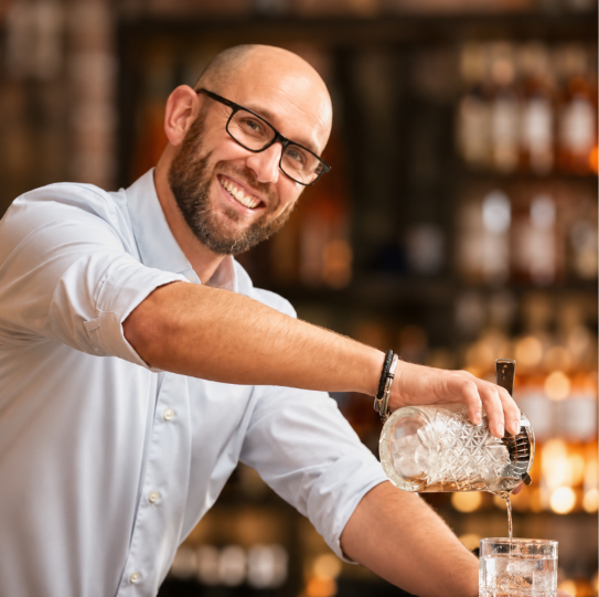Smiling bartender pouring a drink into a glass in a bar setting.