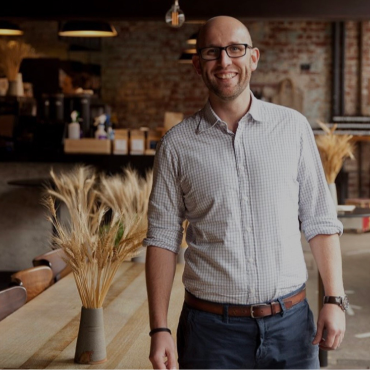 A smiling man with glasses and a checked shirt standing in a cozy cafe with a rustic brick wall and wooden furniture, surrounded by dried wheat in vases.