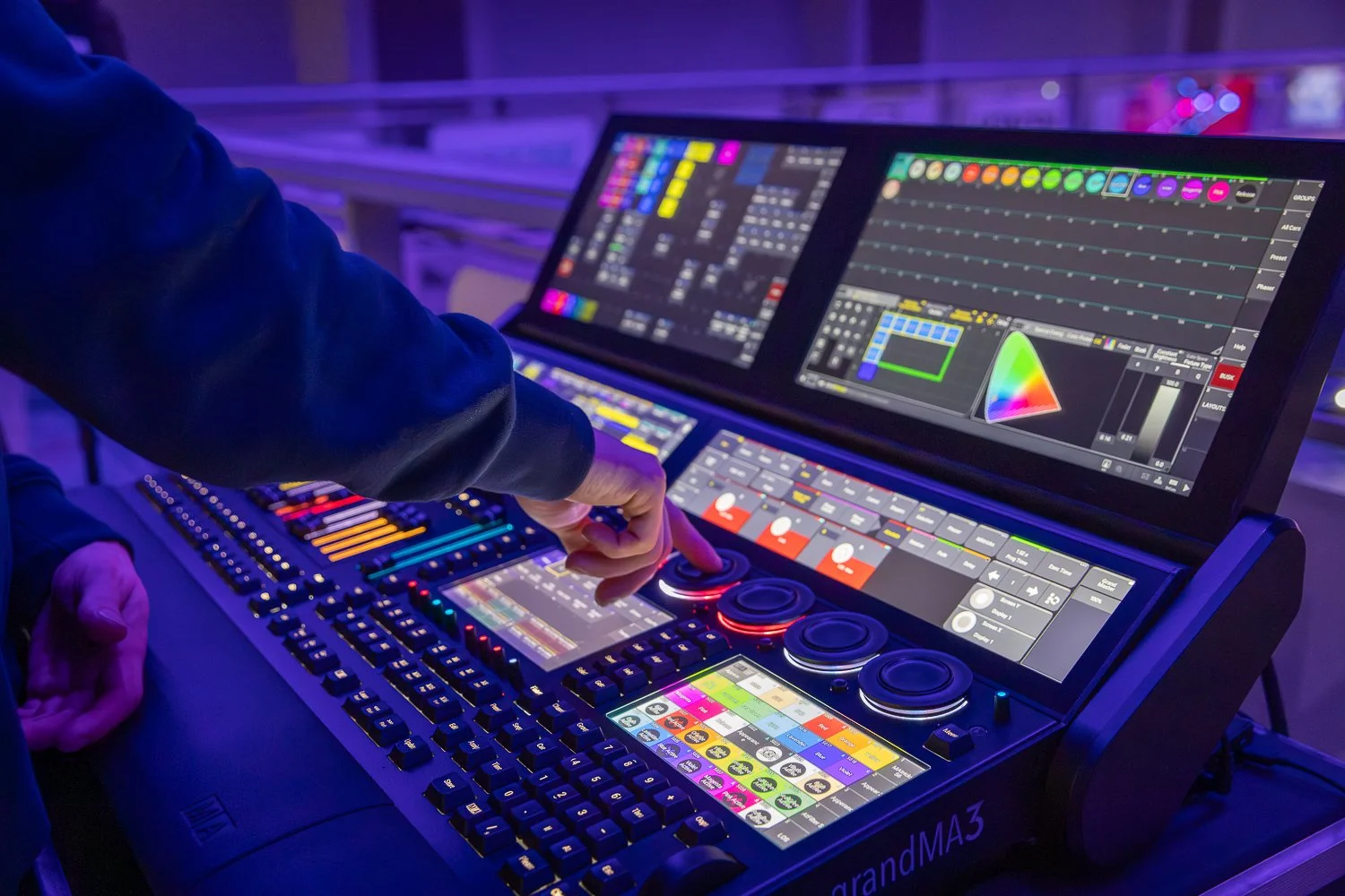 A DJ or music producer using a digital mixing console with multiple screens displaying audio and lighting control software in a dimly lit environment.