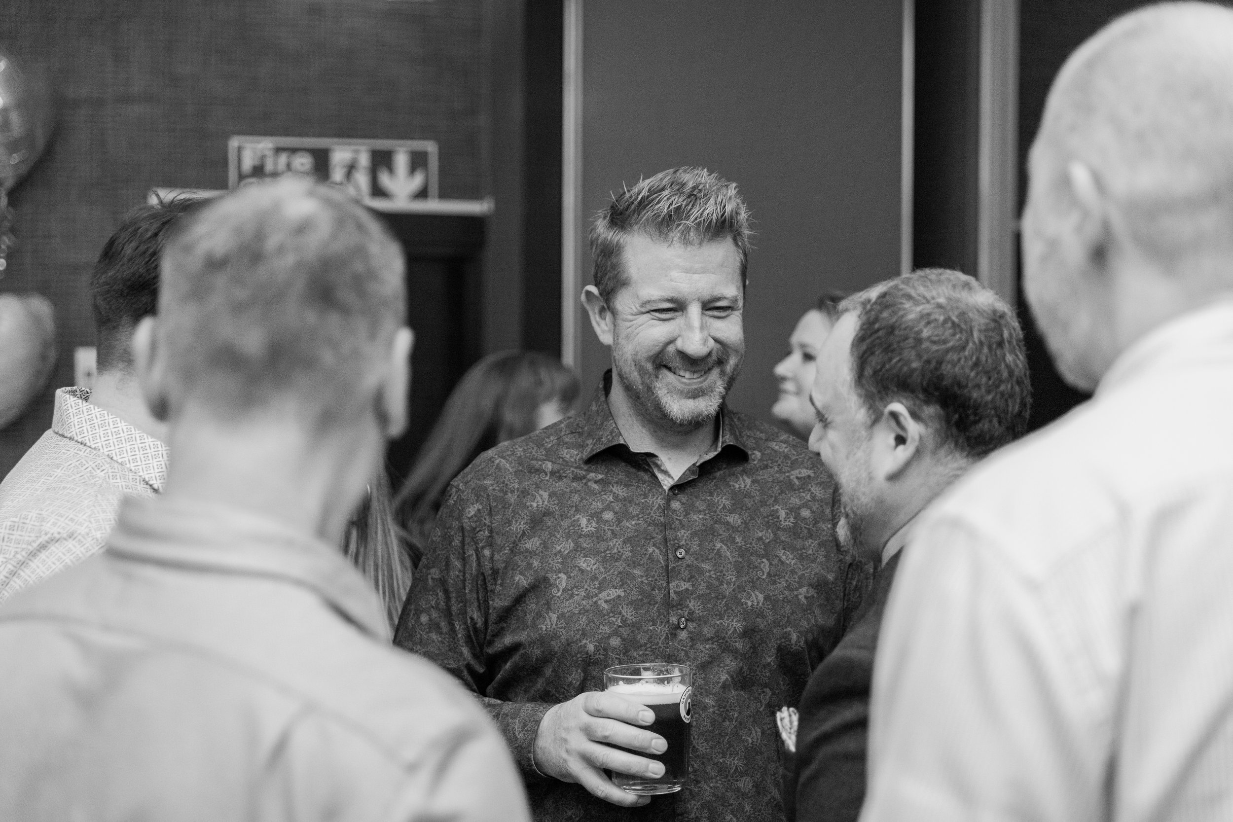 A group of people socializing at a party, with a man in the center holding a glass of dark beer, smiling and engaging in conversation with others.
