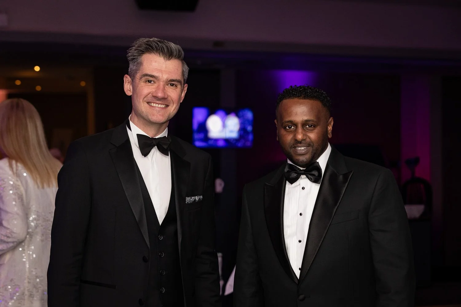 Two men in tuxedos, one with gray hair and the other with black hair, smiling at an event with dim lighting and purple background.
