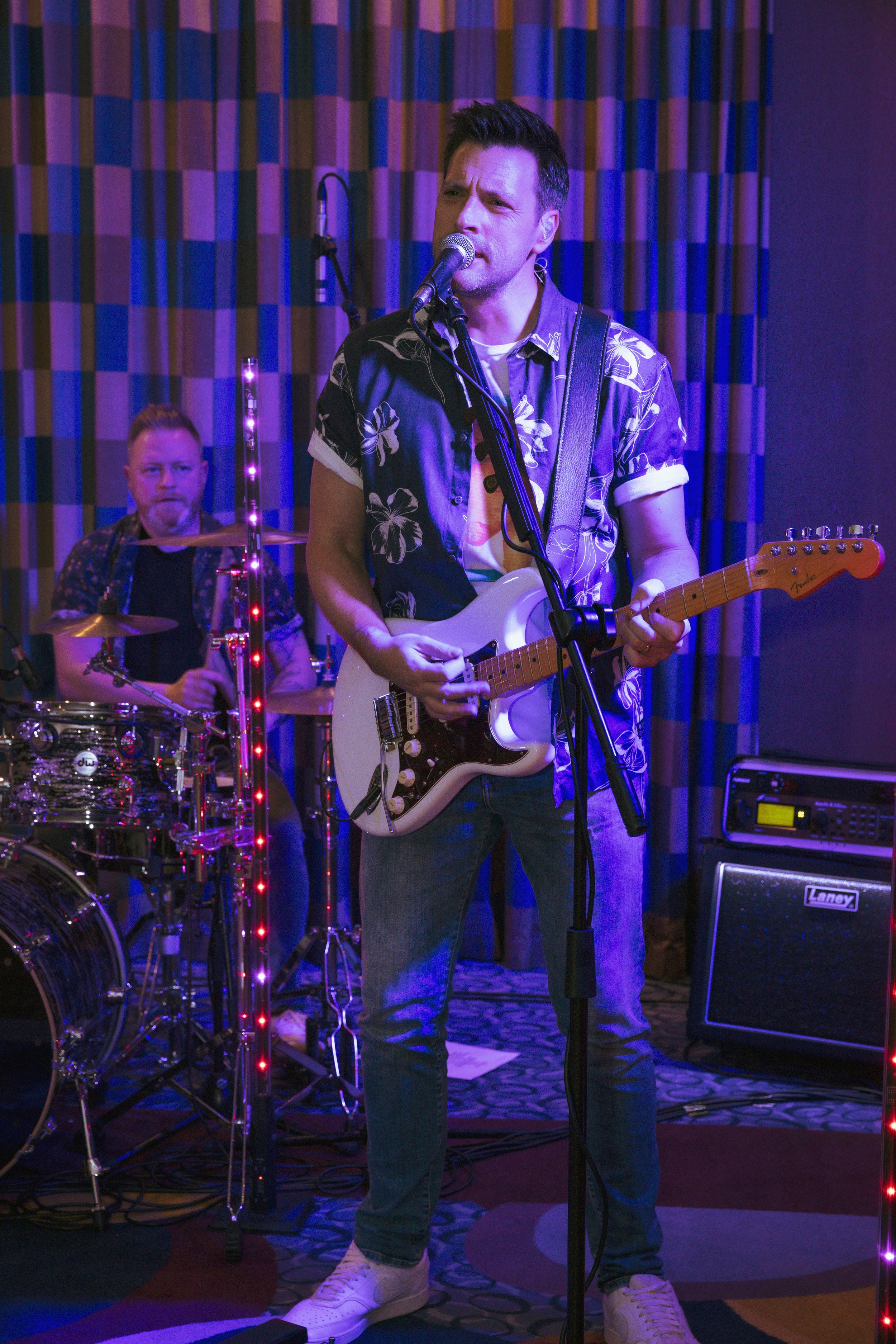Musicians performing on stage with one playing electric guitar and singing into a microphone, the other playing drums in the background, with colorful stage lighting and curtains.