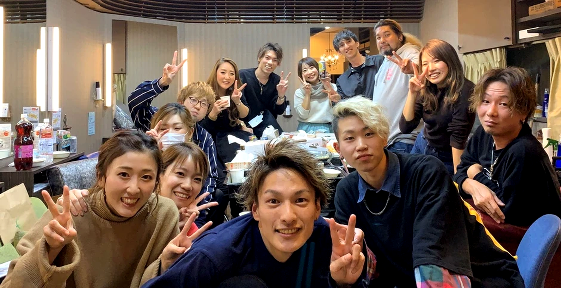 室内でグループ写真を撮っている若者と中年の人の大人数のグループ。皆笑顔でピースサインをしている。