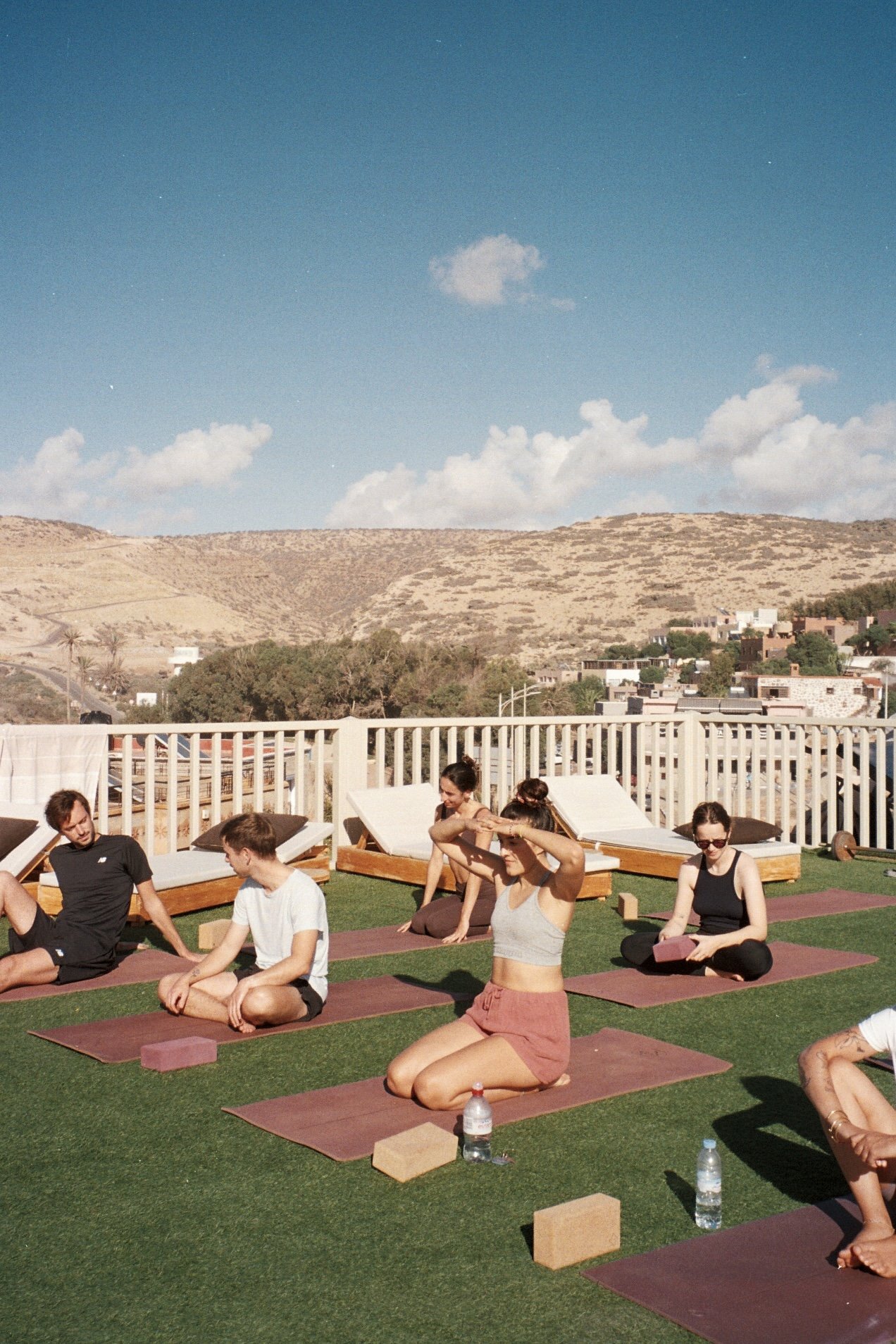Groupe de personnes faisant du yoga ou de la méditation sur un toit avec vue sur une vallée montagneuse en arrière-plan, en position de méditation ou d'étirement, avec des tapis et des blocs de yoga.
