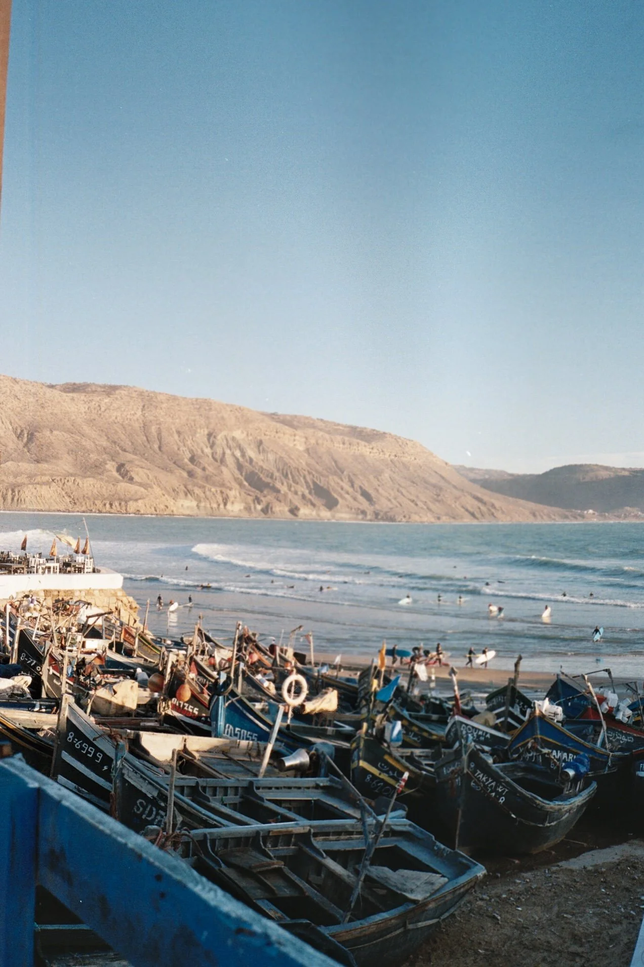 Bateaux de pêche amarrés sur une plage avec des montagnes en arrière-plan et des surfeurs dans l'océan.