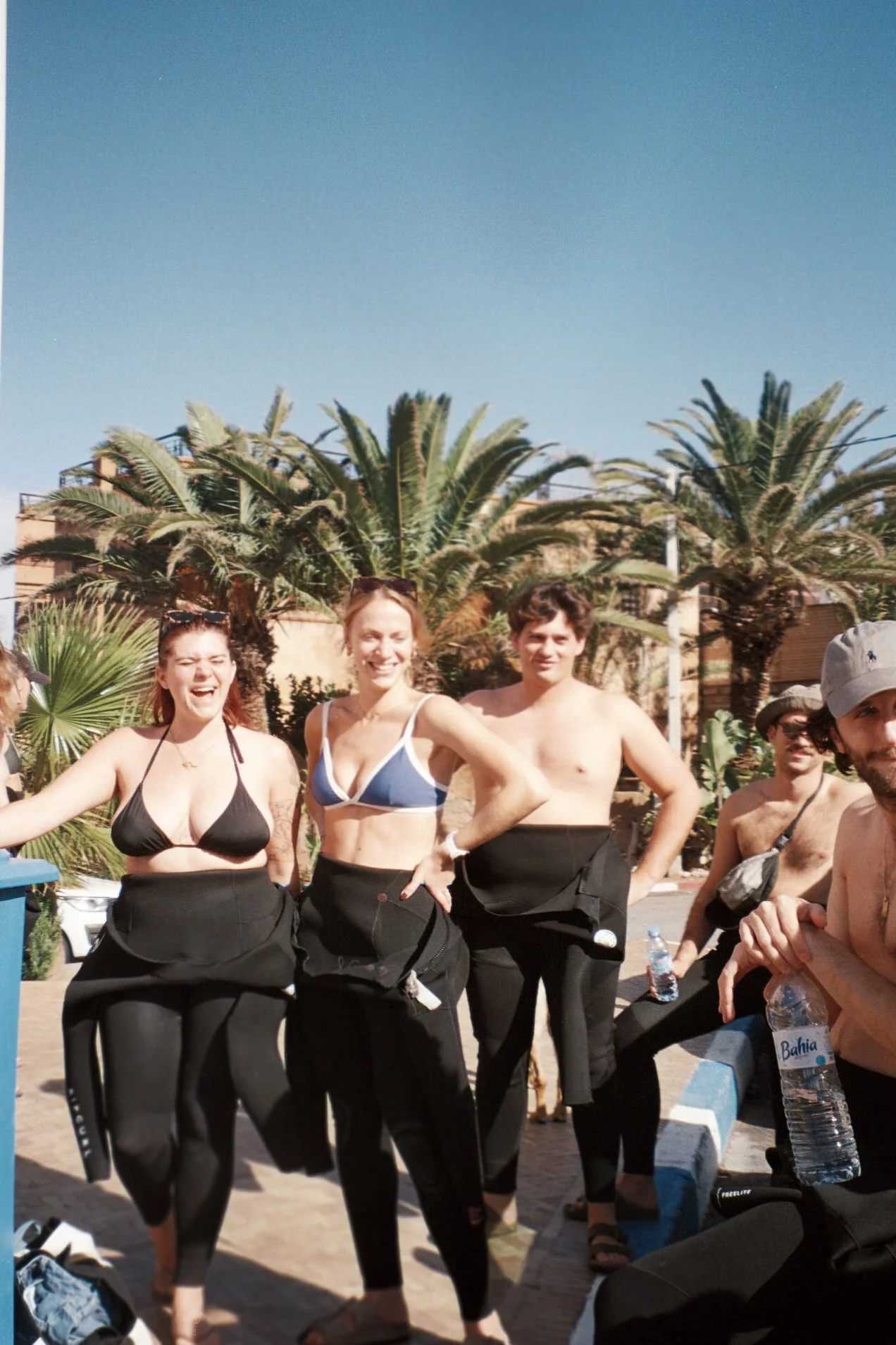 Groupe de personnes en maillots de bain ou semi-nus, souriantes sous un ciel ensoleillé avec des palmiers en arrière-plan.