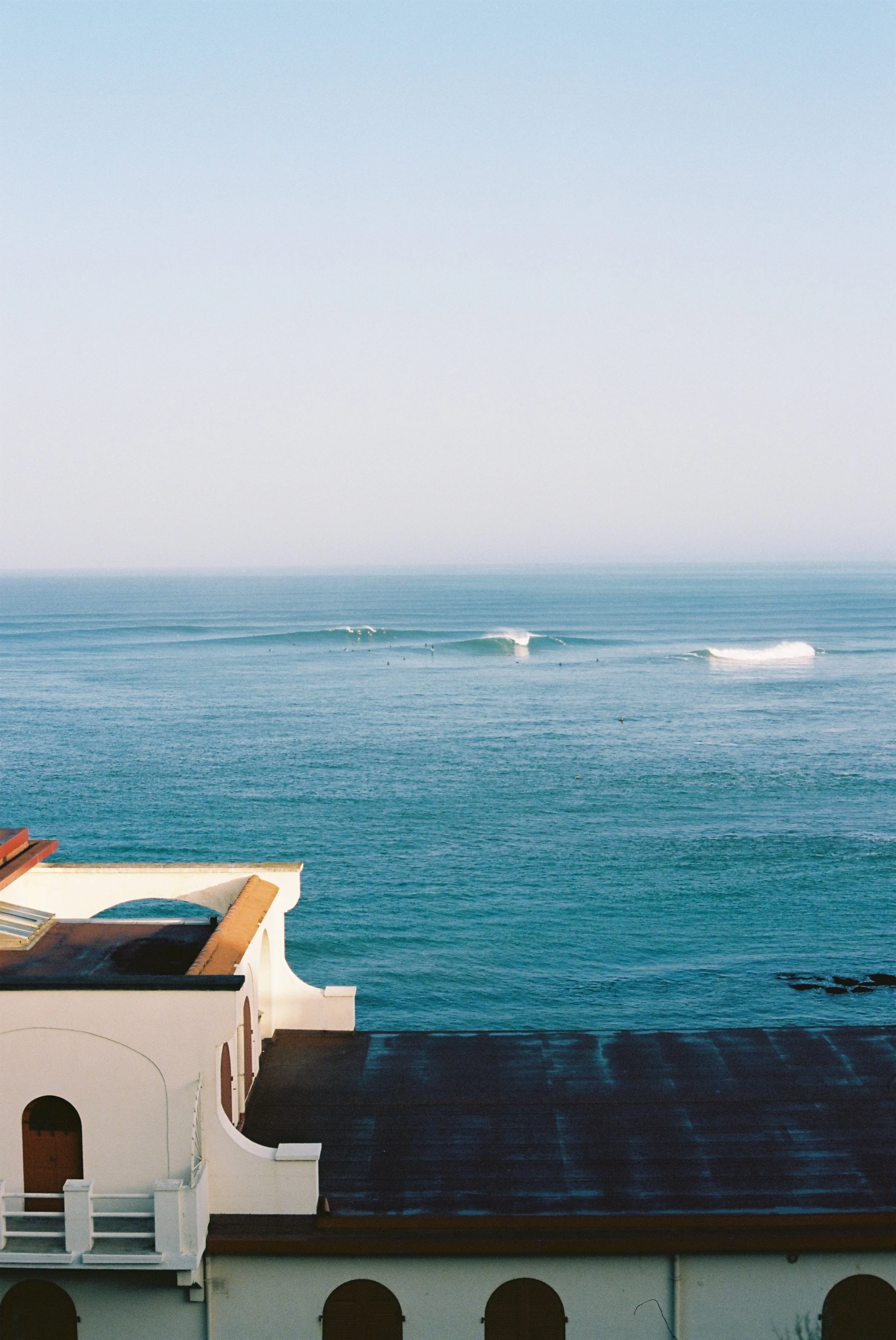 Vue de l'océan avec des vagues, vue partielle d'un bâtiment blanc avec un toit en bois, au premier plan.