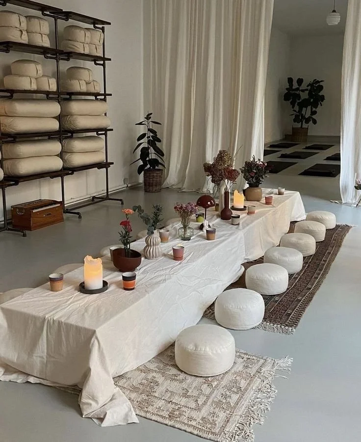 Une table longue couverte d'une nappe blanche, décorée de fleurs, bougies et vaisselle, entourée de coussins arrondis, dans une pièce lumineuse avec des étagères de linge et des plantes d'intérieur.