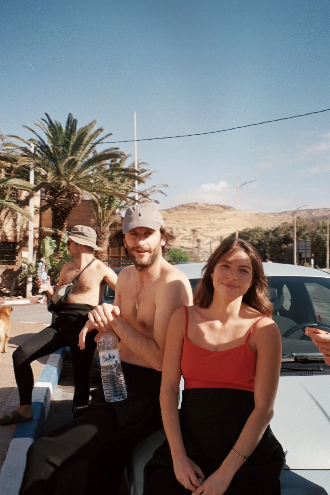Deux jeunes adultes, un homme torse nu portant un chapeau et une femme portant une robe rouge, posent et sourient devant une voiture en plein jour avec des palmiers et des montagnes en arrière-plan.