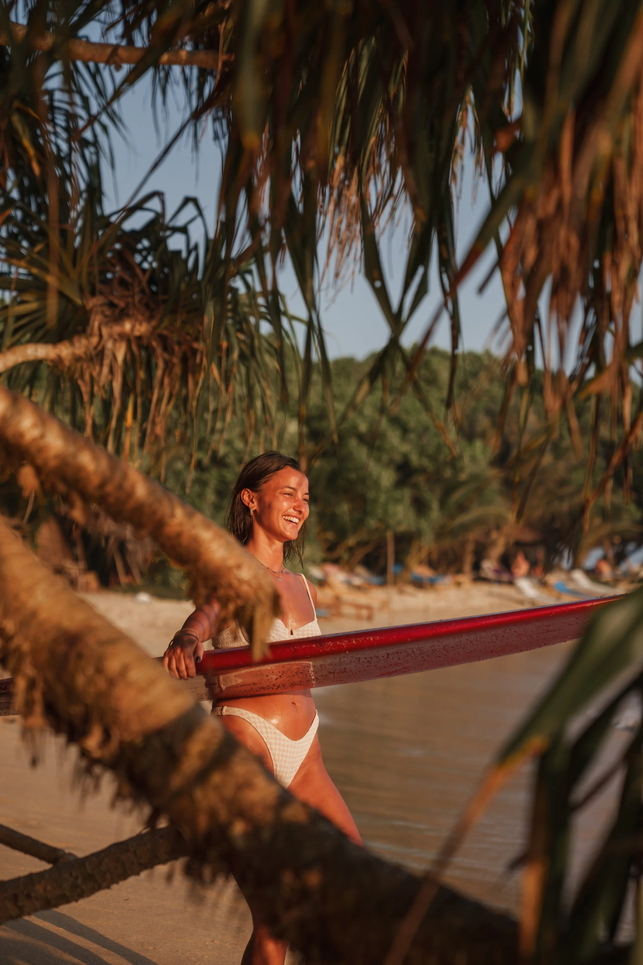 Une femme souriante en maillot de bain, tenant une planche de surf rouge, sur une plage en fin d'après-midi ou coucher de soleil, entourée de végétation tropicale.