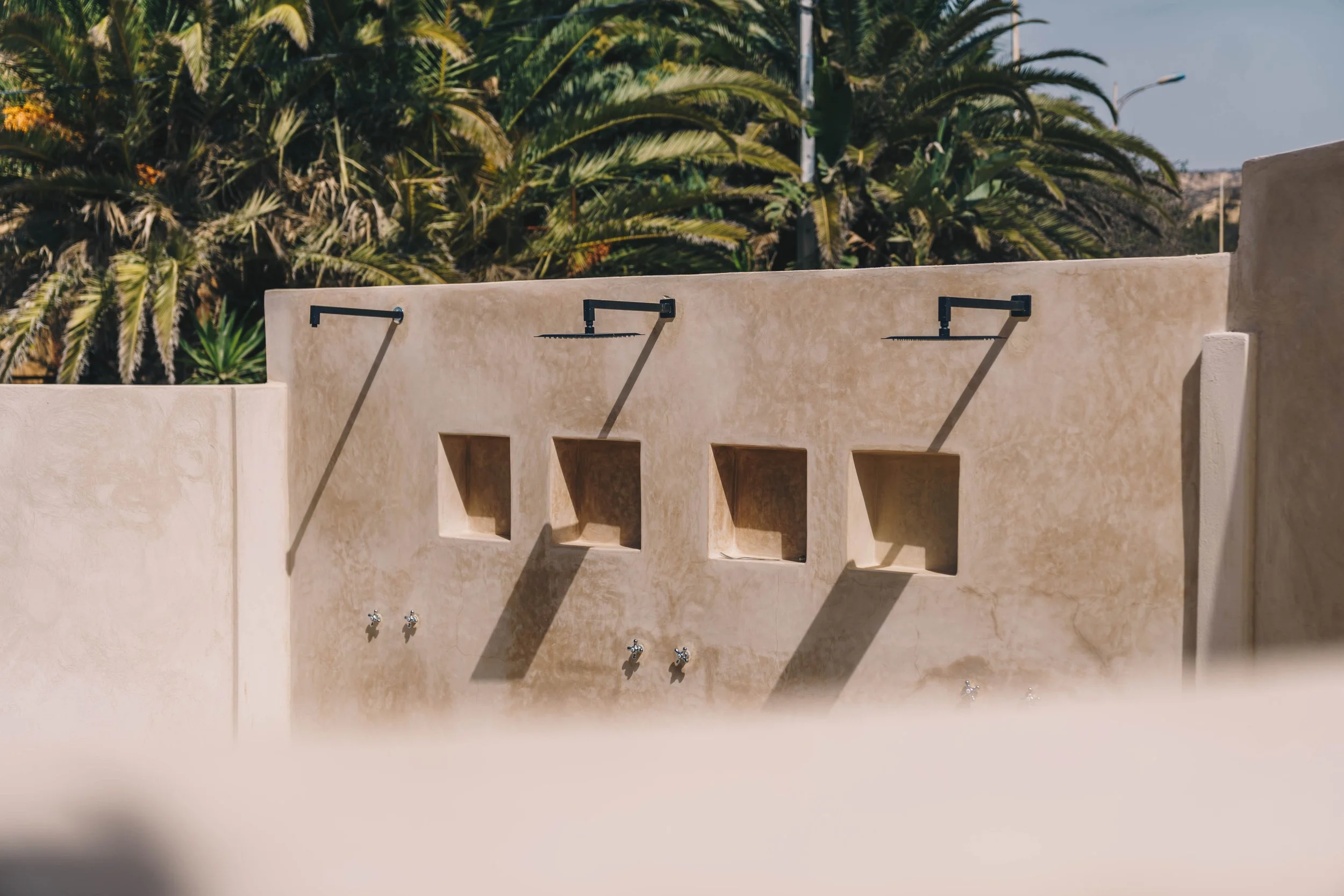 Douze bornes de lavage en extérieur avec des robinets, construite en béton beige, avec des ombres projetées par le soleil, et des palmiers en arrière-plan.