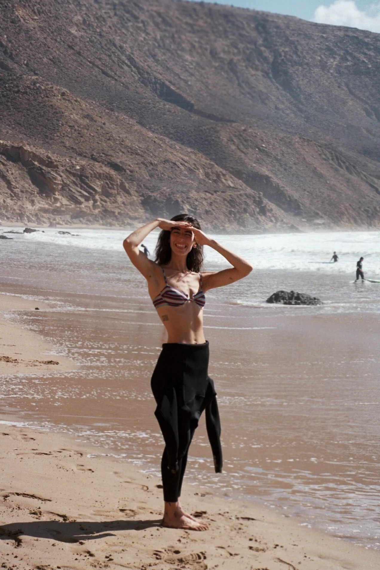 Une femme sourire sur une plage, en train de regarder vers le soleil en portant un maillot de bain rayé et un pantalon noir noué à la taille, avec des montagnes en arrière-plan.