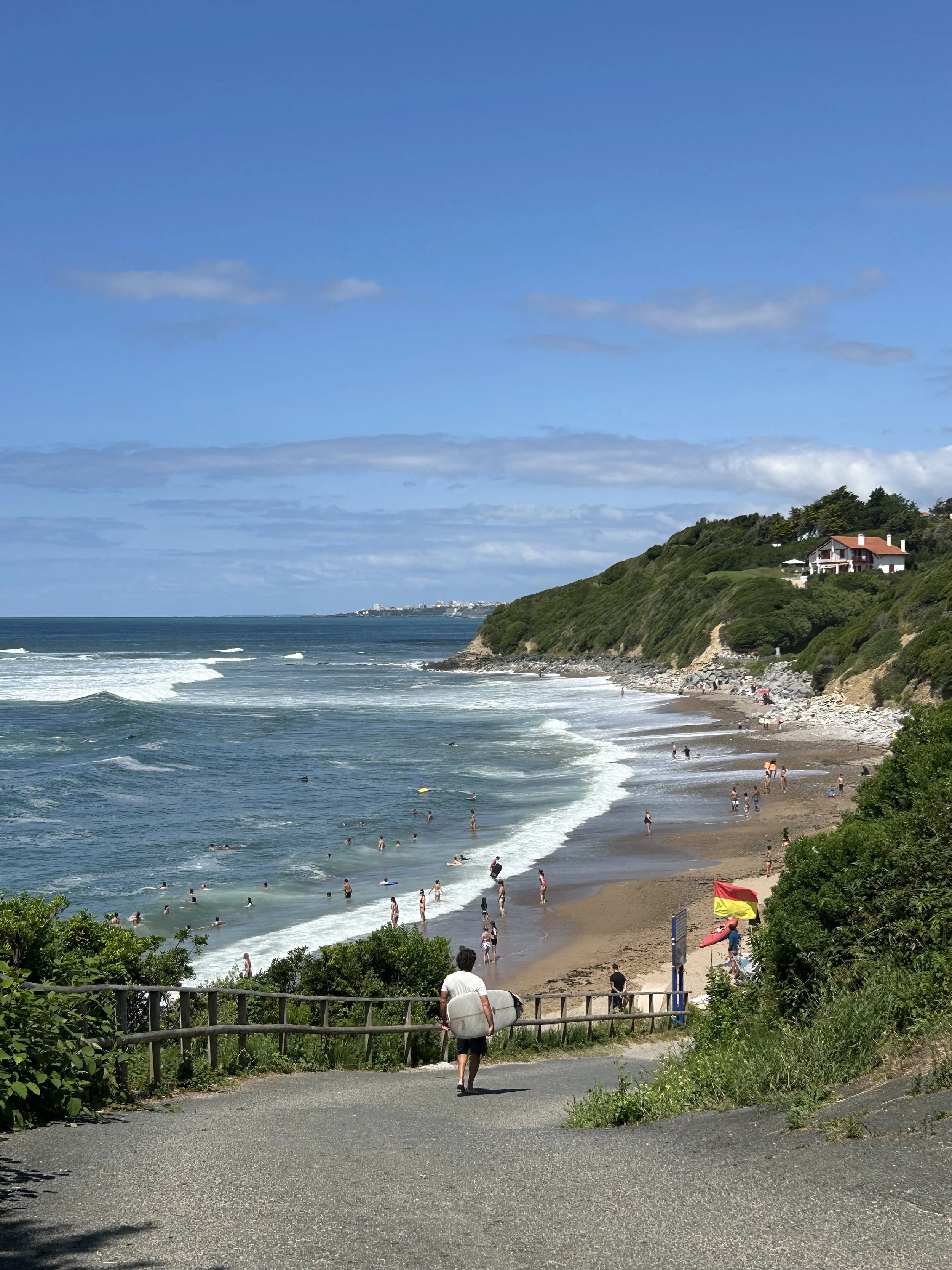 Plage en bord de mer avec des baigneurs, un promeneur avec une planche de surf, falaises verdoyantes et maisons en arrière-plan, ciel bleu avec quelques nuages.