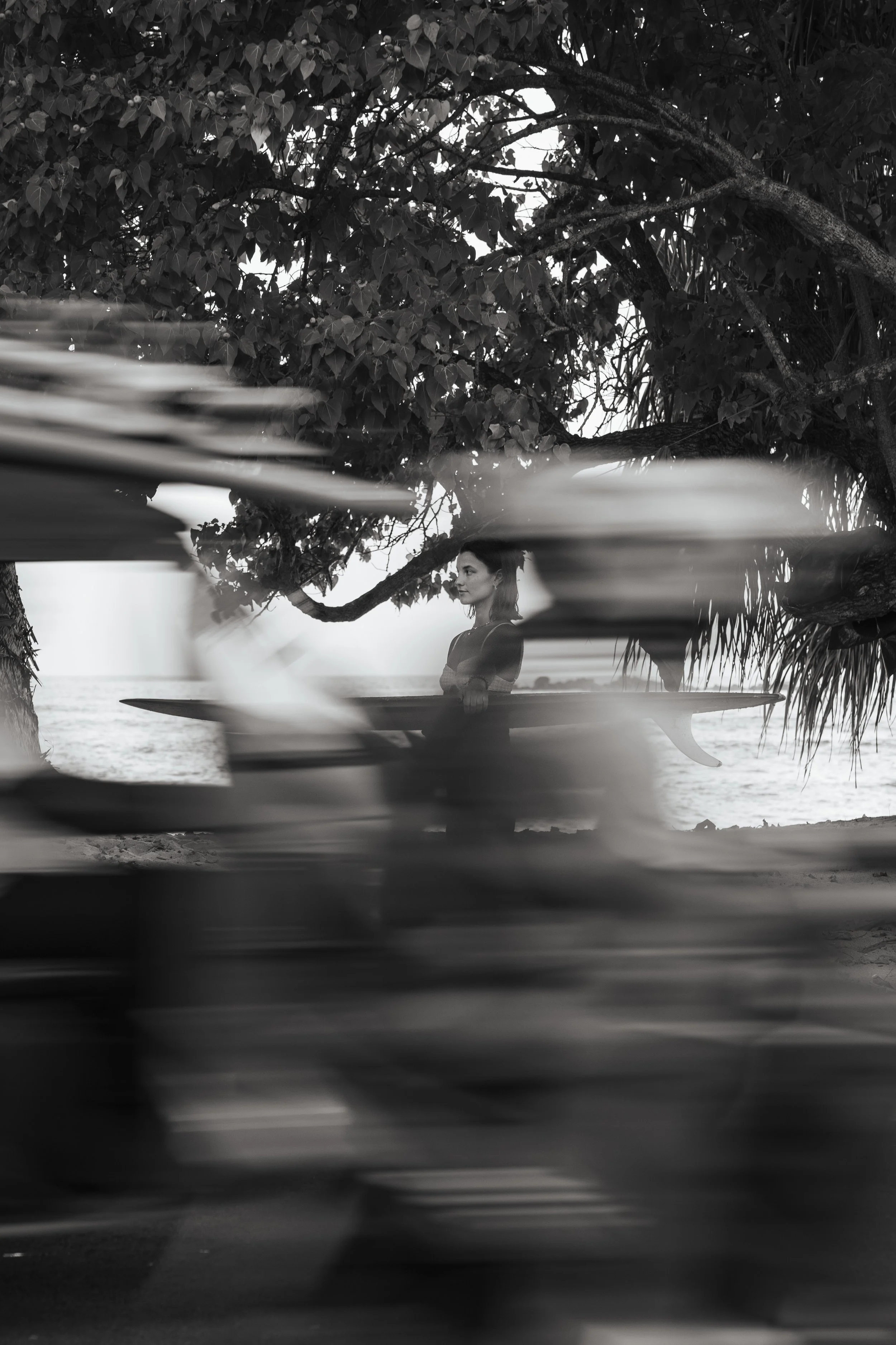 Une femme avec une planche de surf, vue en contre-plongée à travers des branches floues, sur une plage proche de l'océan, en noir et blanc.