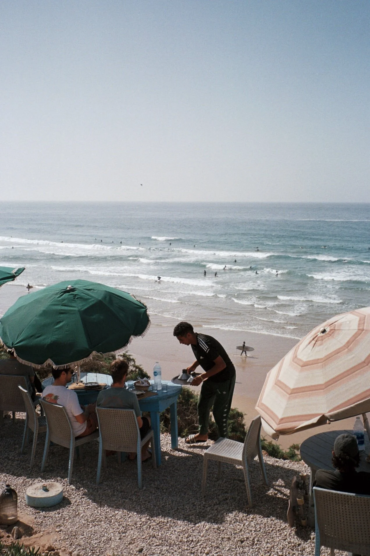 Des personnes profitent d'une vue sur la plage depuis une terrasse avec parasols, quelques surfeurs dans l'eau, et un garçon servant de la nourriture ou des boissons à une table.