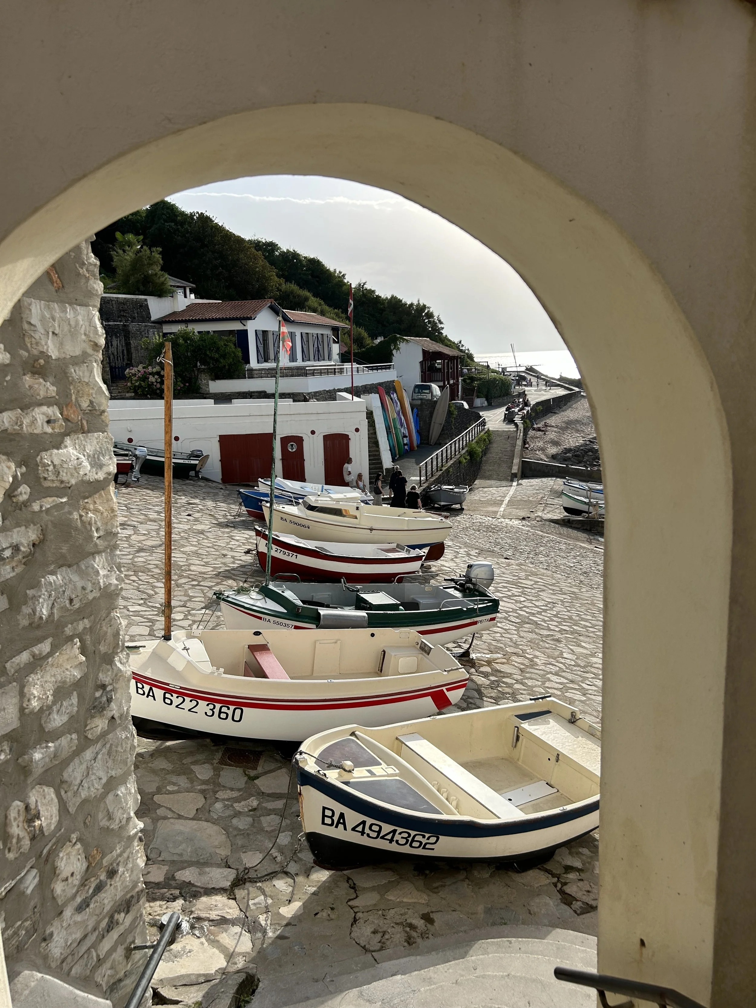 Petits bateaux de pêche alignés sur une voie pavée près de la mer, vue à travers une arche, avec des maisons et des kayaks en arrière-plan.